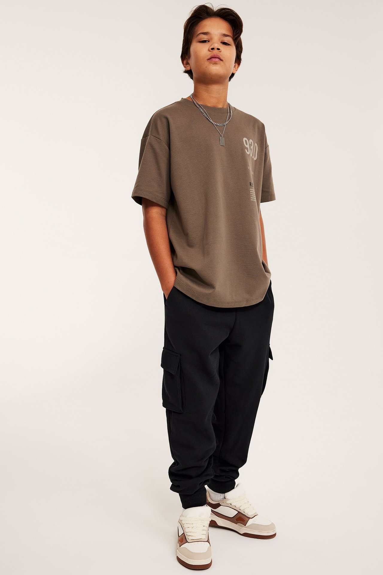 Widok z przodu: chłopiec w czarnych dresach cargo, brązowej koszulce i białych trampkach.