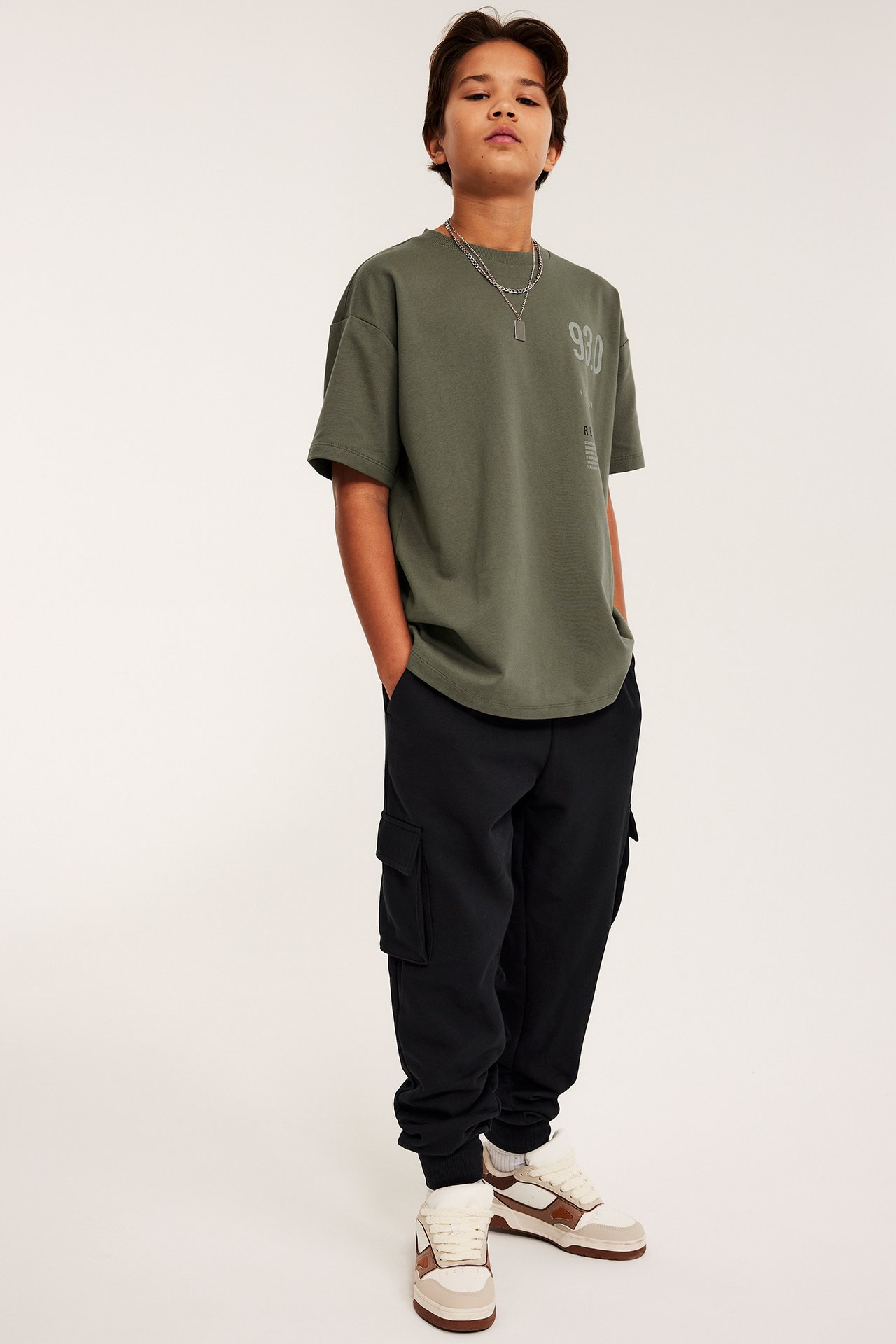 Edestä: Poika mustissa cargolenkkihousuissa, oliivinvihreässä t-paidassa ja lenkkikengissä.