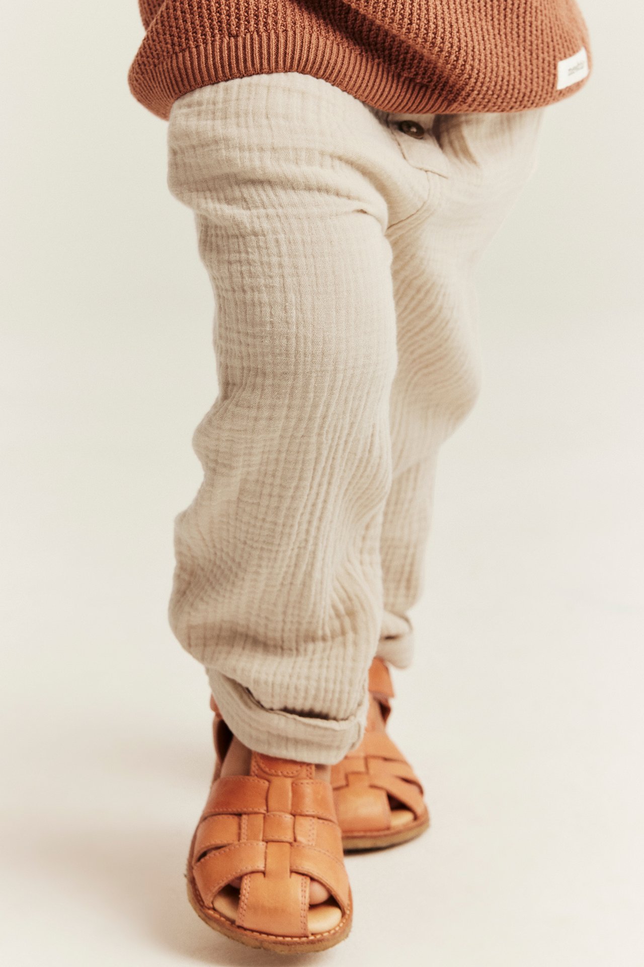 Edestäpäin: vauva yllään luonnonvalkoiset puuvillamusliinihousut käärityillä lahkeilla, ruskea neulepaita ja ruskeat sandaalit.