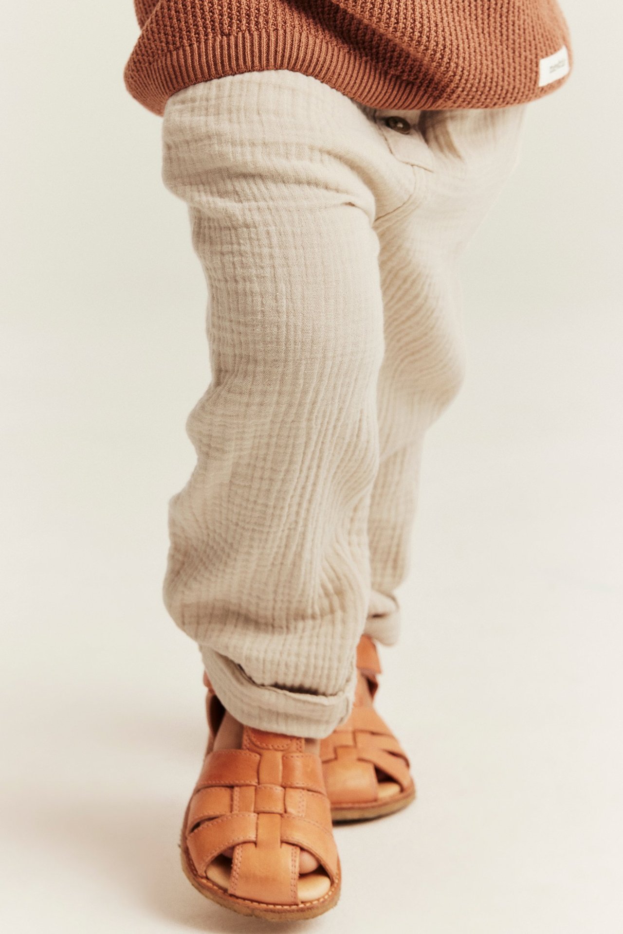 Forfra: baby i kremhvite bomullsmuslinbukser med oppbrettede ben, brun strikket topp og brune vevde sandaler.