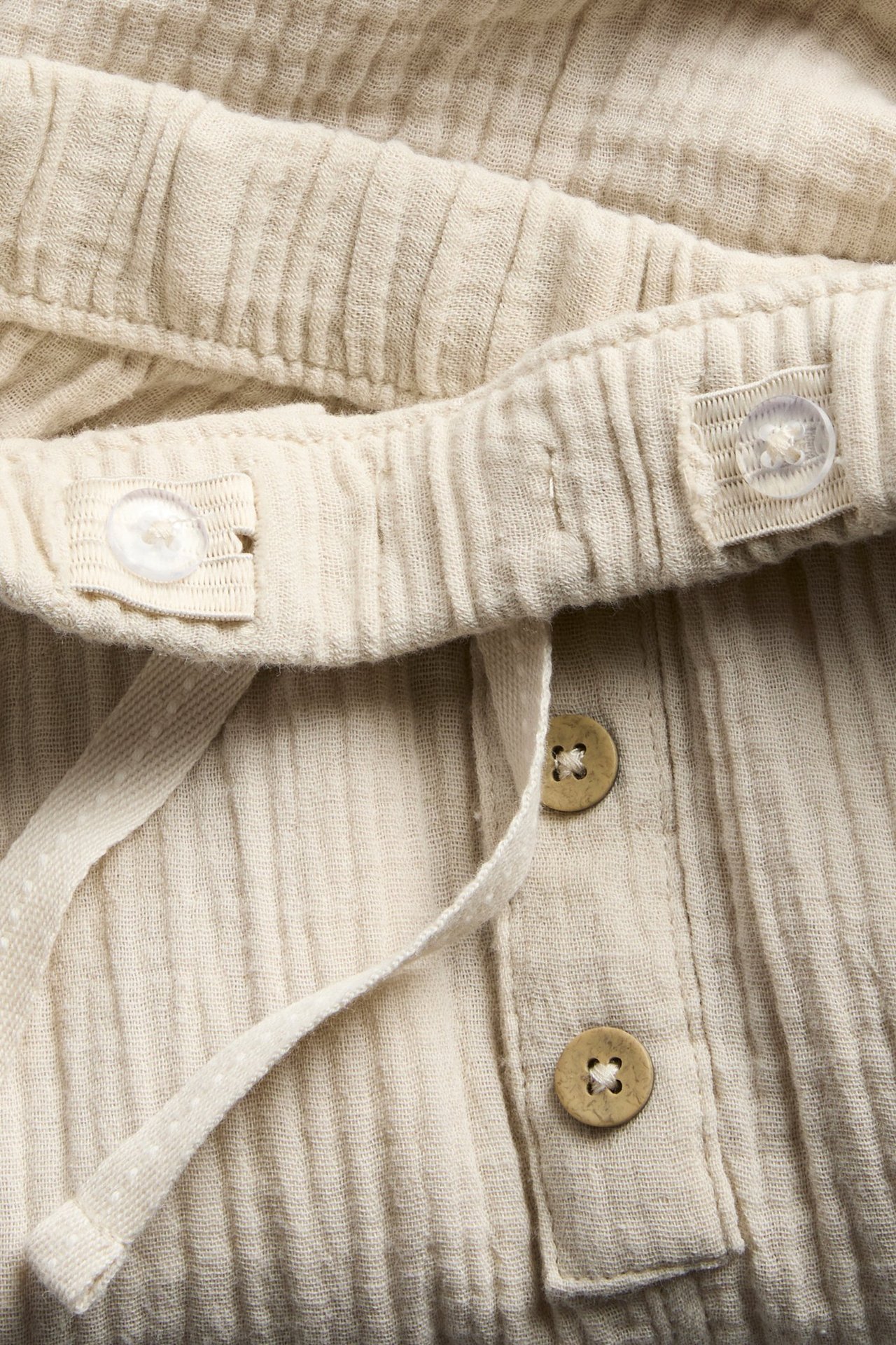 Nærbilde av offwhite bomullsmuslinbukse med justerbar elastisk midje, dekorativ snøring og knapper foran.