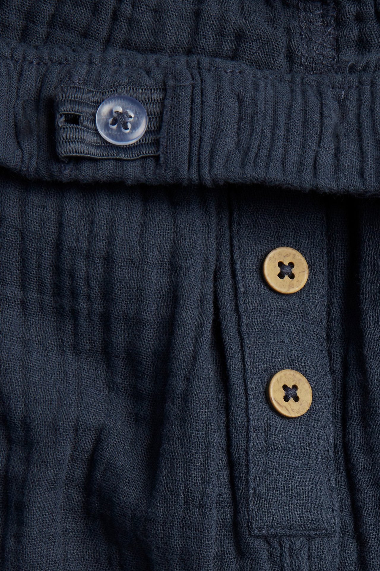 Lähikuva tummansinisistä puuvillamusliinihousuista, joissa säädettävä vyötärönappi ja kaksi koristepainiketta edessä.