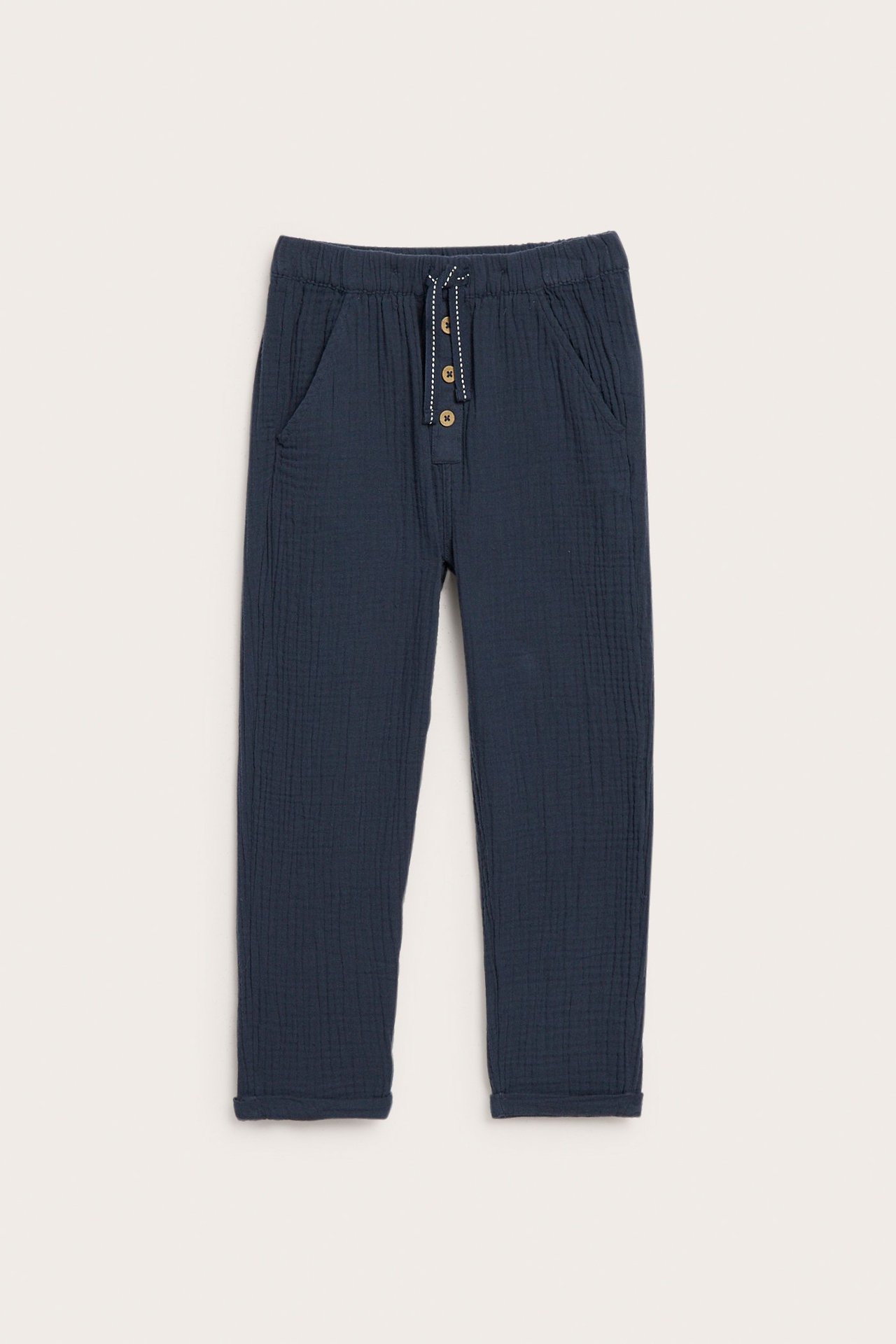 Widok z przodu: ciemnoniebieskie bawełniane spodnie dziecięce z muślinu, elastyczny pas, sznurek, guziki, podwinięte nogawki.