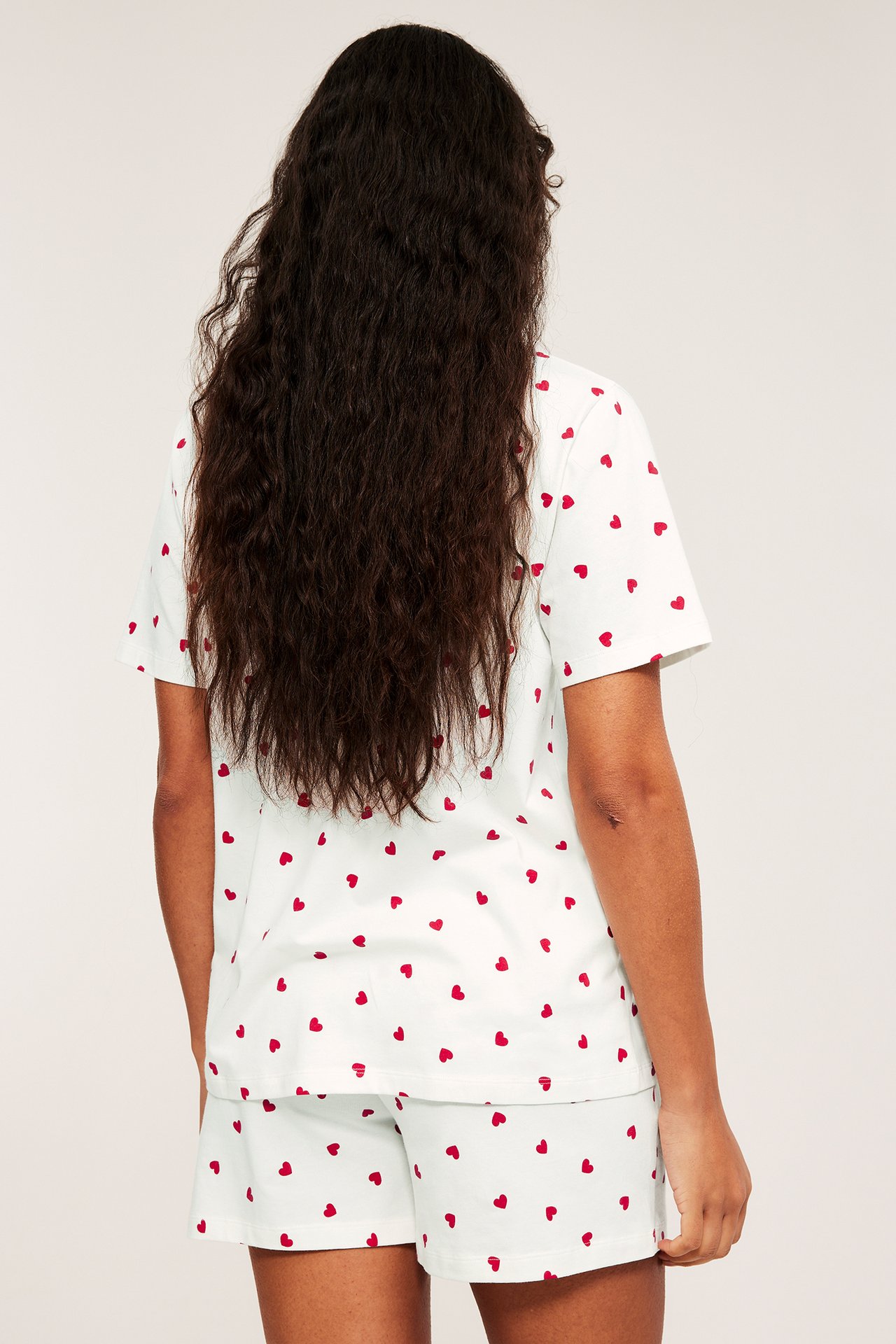 Bakfra: Kvinne i offwhite jersey-pyjamas med rødt hjerteprint, kortermet topp og shorts.