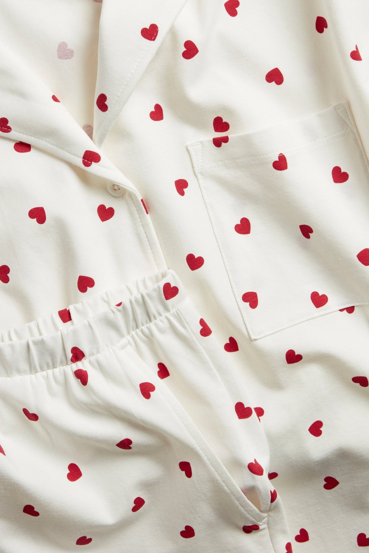 Närbild av offwhite trikåpyjamas med rött hjärtmönster. Toppen har krage, shorts har resår i midjan.