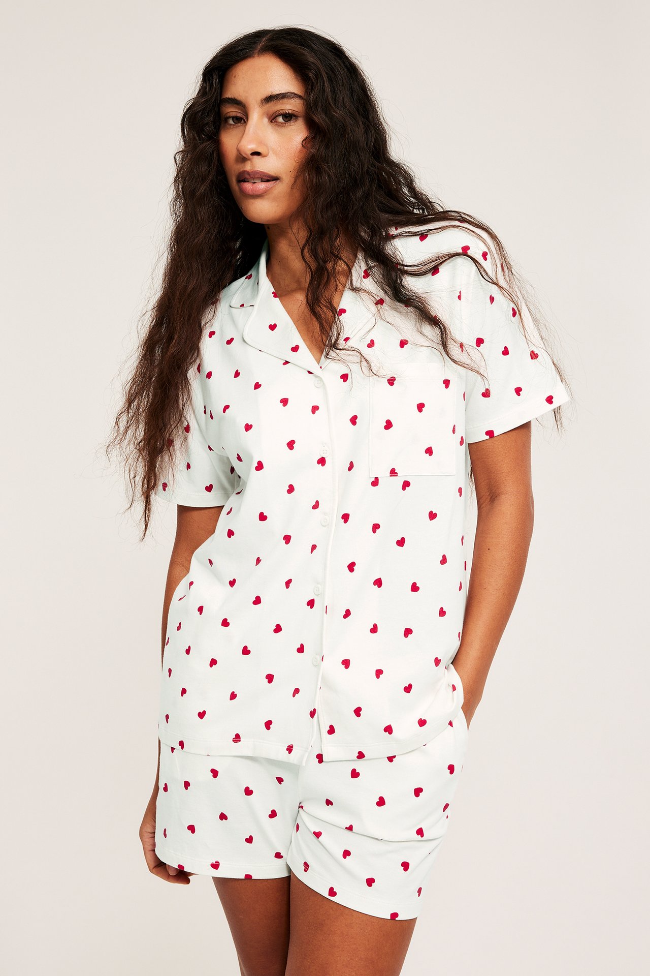 Edestäpäin, nainen yllään luonnonvalkoinen trikoopyjama punaisilla sydämillä, lyhythihainen yläosa ja taskulliset shortsit.