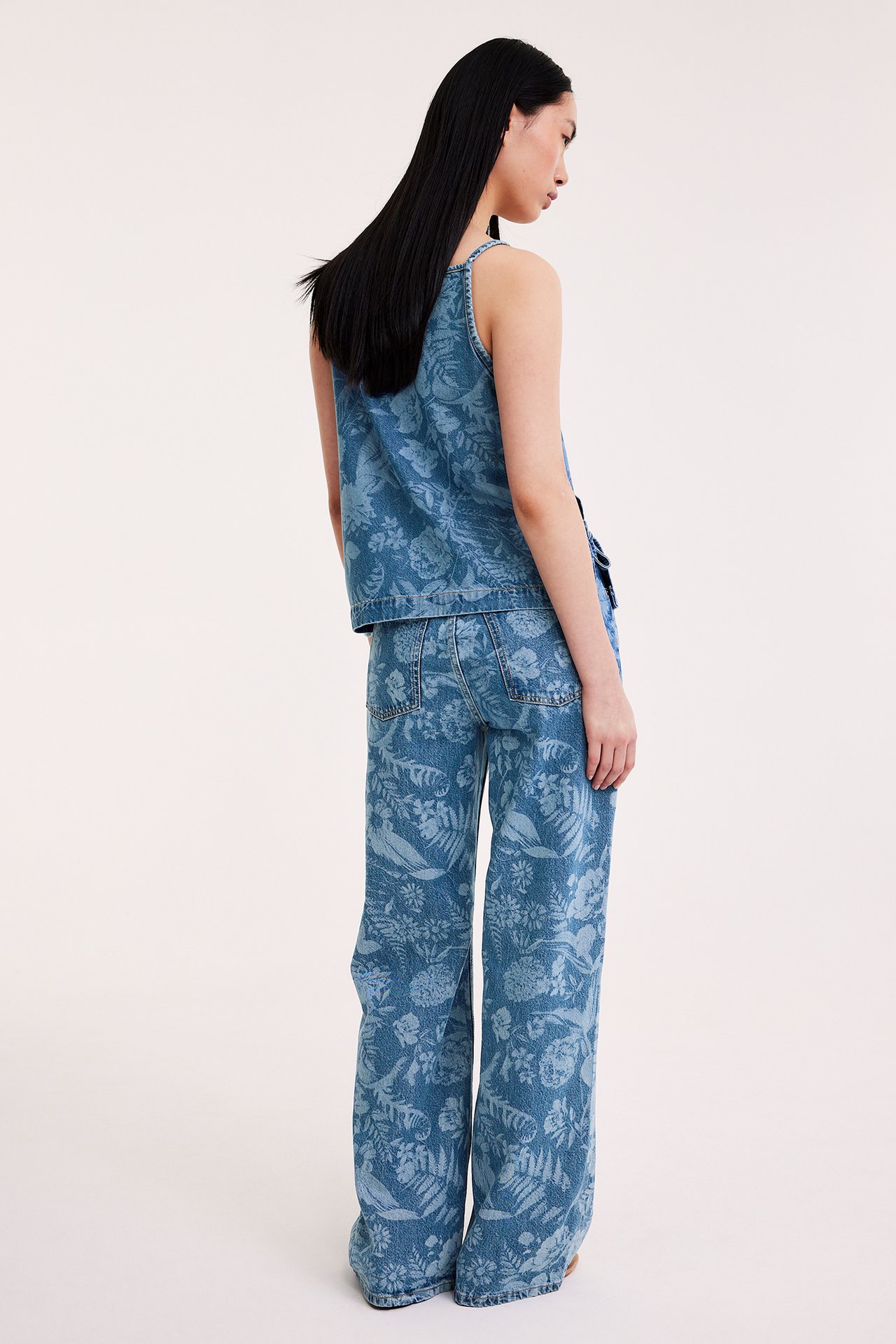 Nainen takaa, yllään vaaleat kukkakuviolliset leveälahkeiset farkut ja samanlainen toppi.