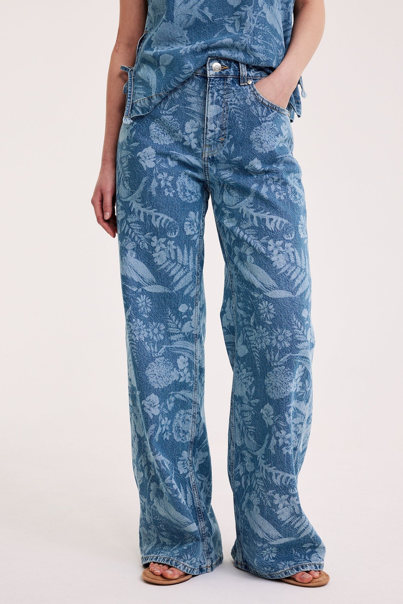 Nainen edestäpäin: yllään vaaleansiniset kukkakuvioiset leveälahkeiset farkut, yhteensopiva toppi ja ruskeat sandaalit.