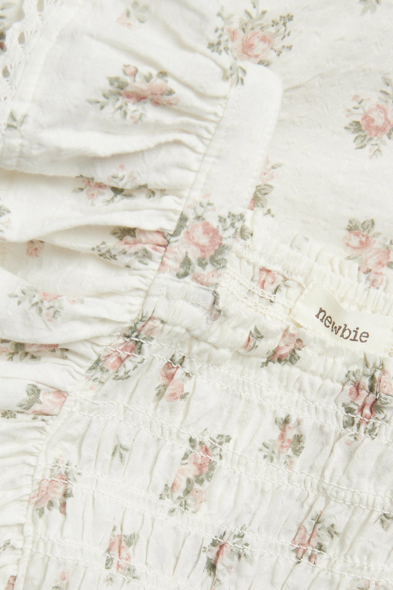 Lähikuva luonnonvalkoisesta kukkakuvioisesta kudotusta haalarista, jossa röyhelöitä, pitsiä ja smock-resori.