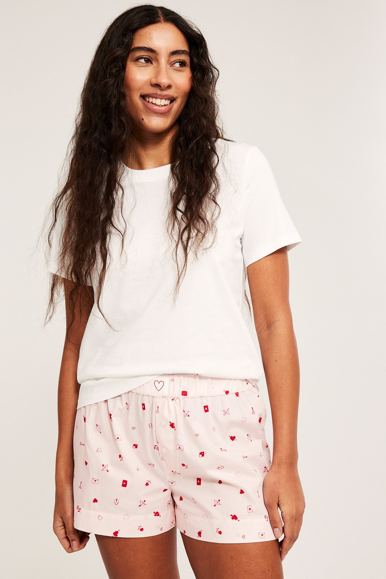 Forfra: kvinne i lyserosa mønstrede pysjamasshorts i bomull med elastisk midje og hvit t-skjorte.