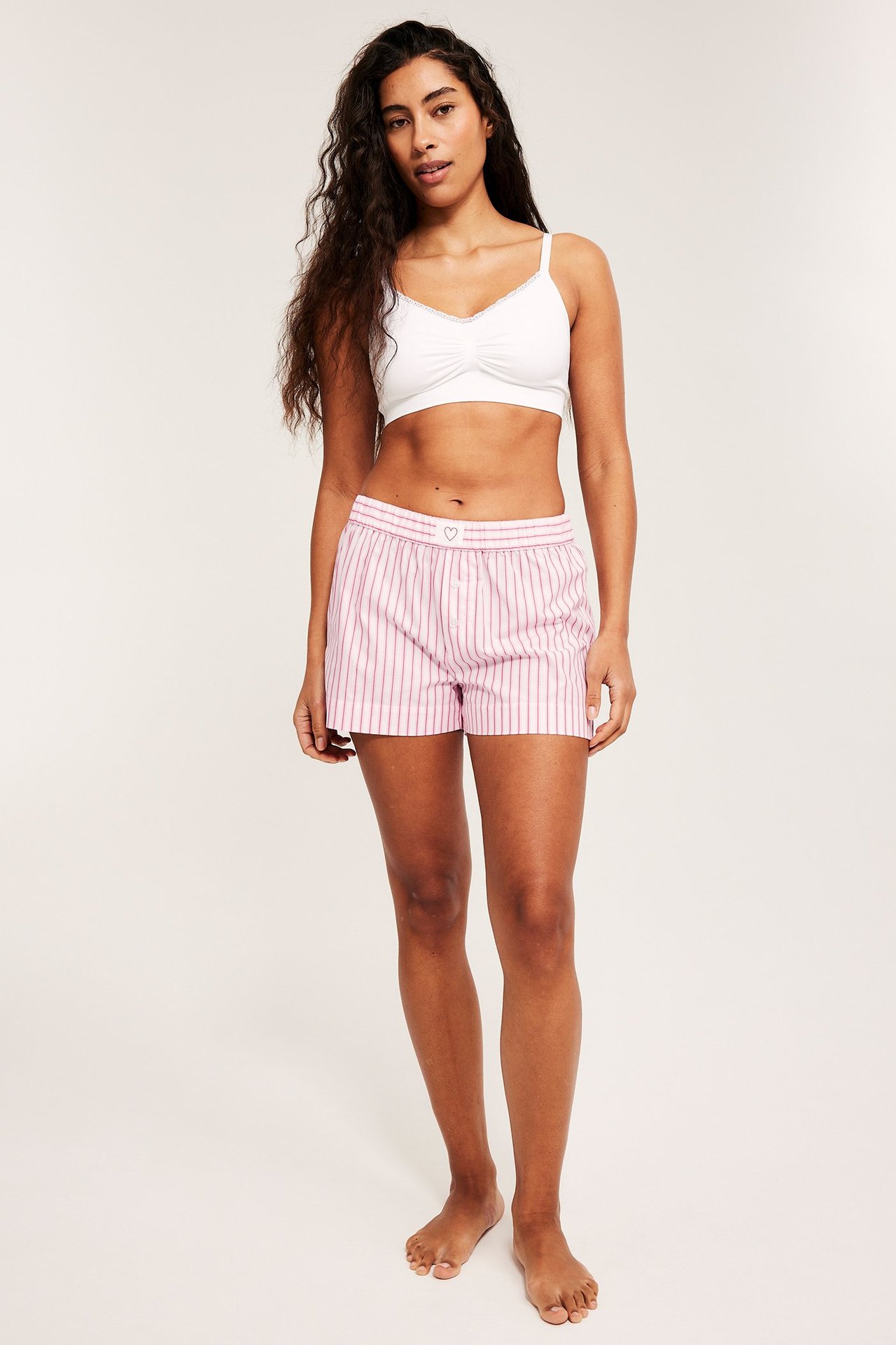 Nainen edestäpäin, yllään vaaleanpunaiset raidalliset pyjamashortsit ja valkoinen bralette.