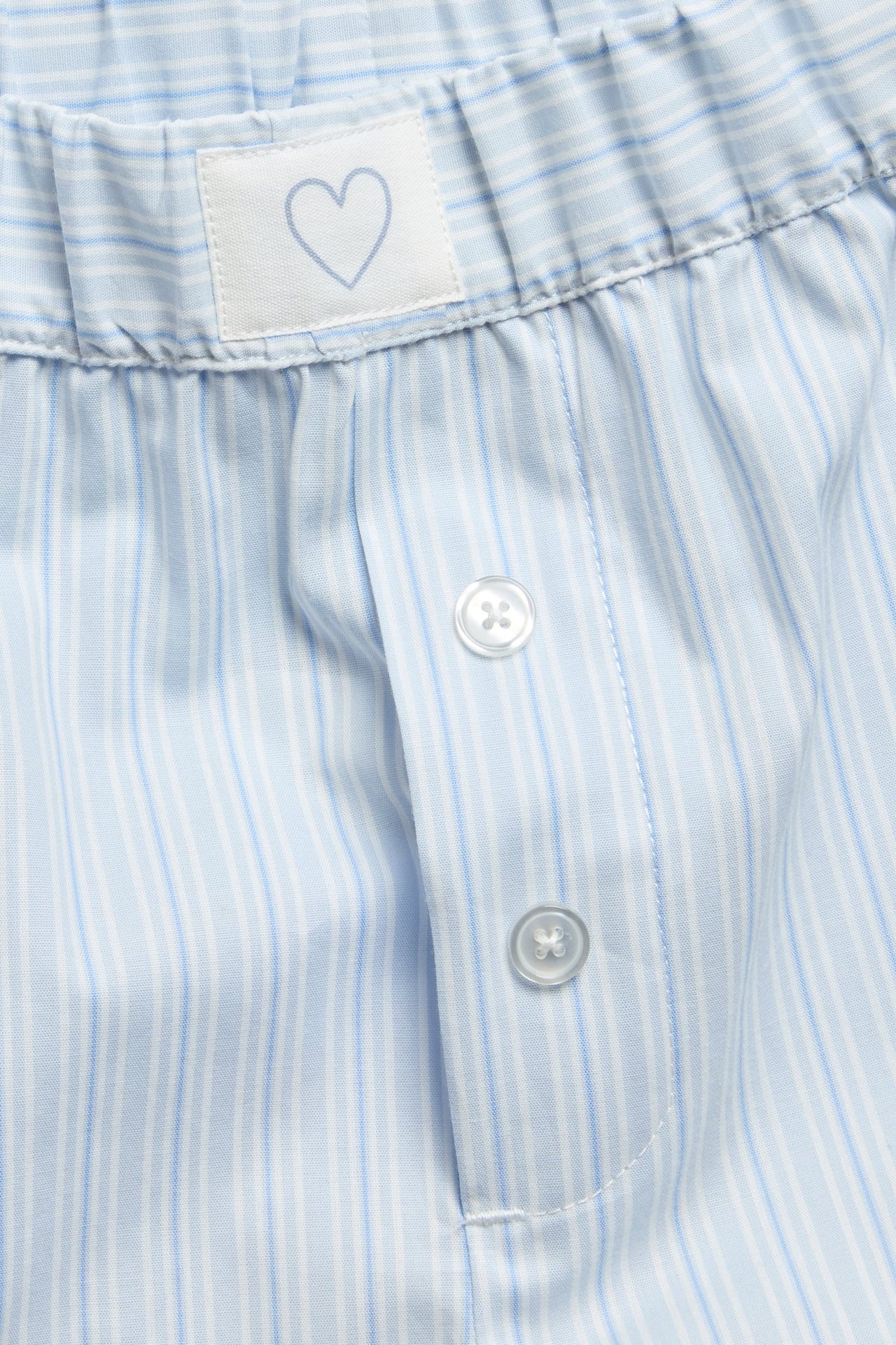 Närbild av blå- och vitrandiga pyjamasshorts i bomull med resår i midjan, dekorknappar och hjärtetikett.