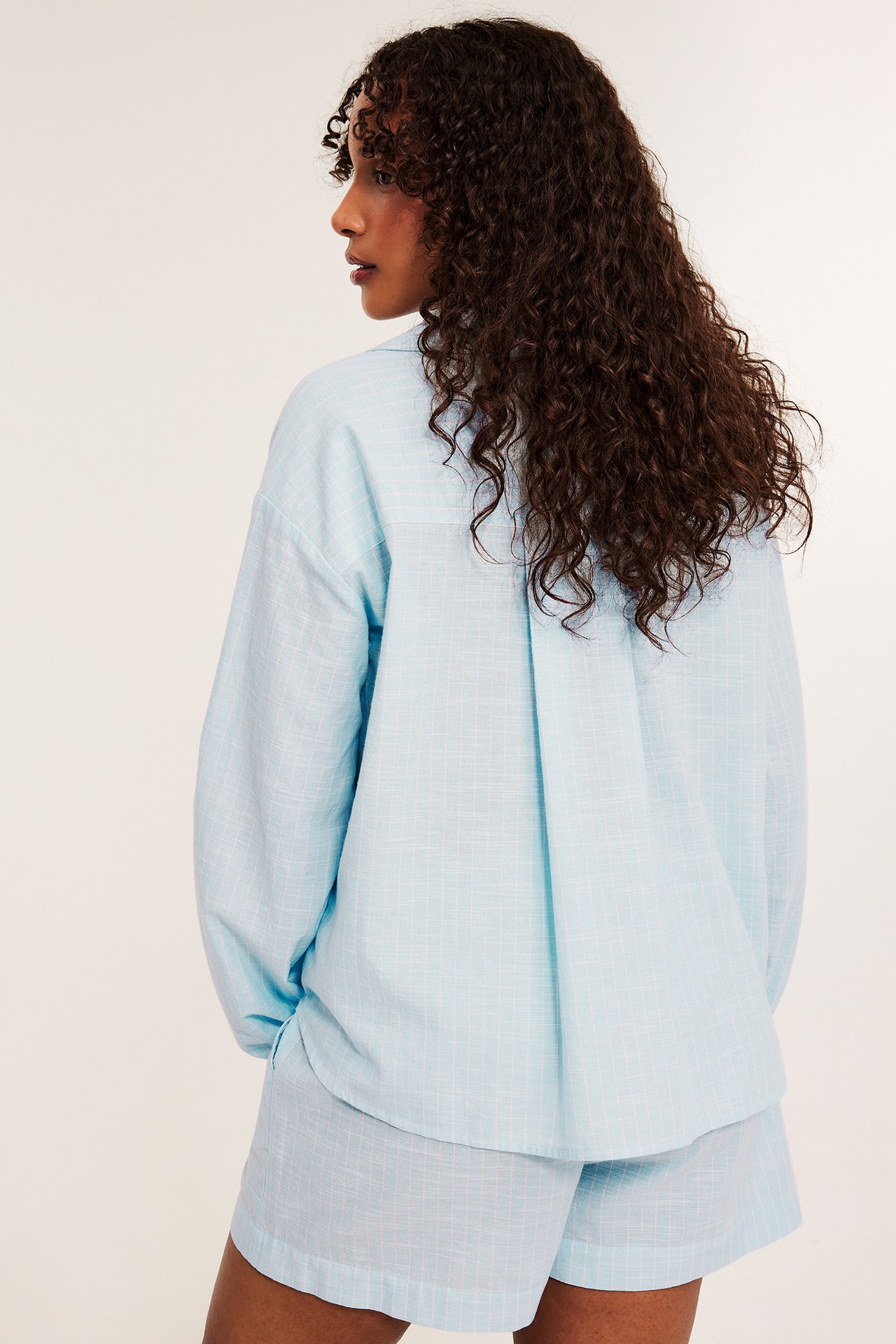 Bakifrån på kvinna i ljusblå randig bomullspyjamasskjorta och matchande shorts.