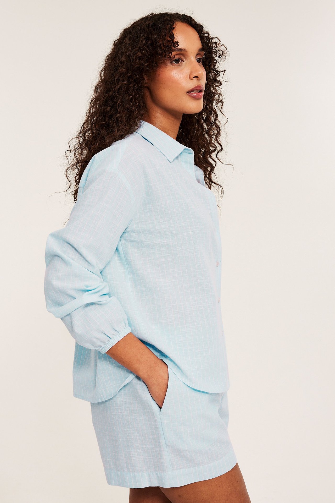 Naisen sivukuva vaaleansinisessä raidallisessa puuvillapyjamassa: löysä paita, shortsit käsi taskussa.