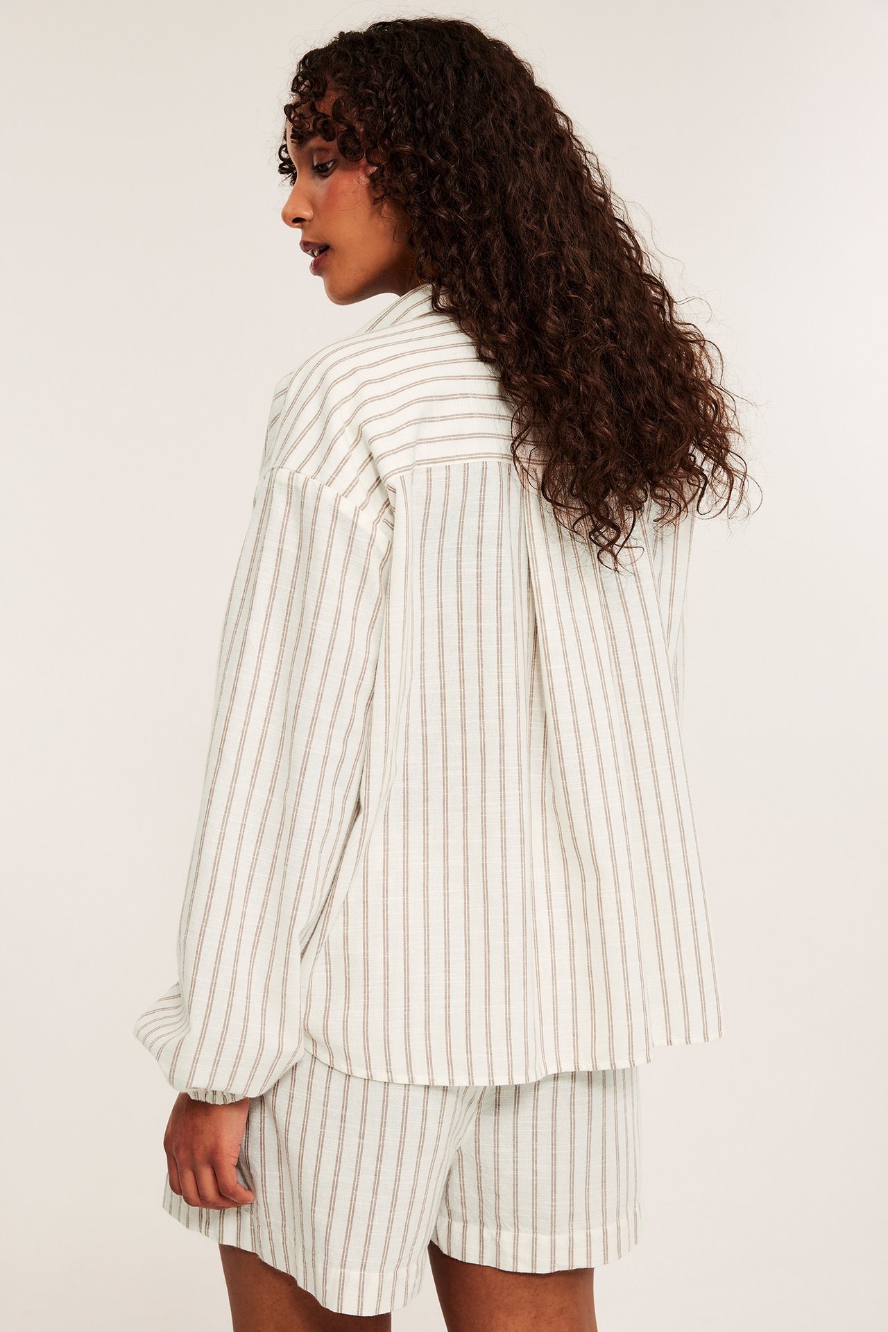 Bakfra: kvinne i et stripete beige pyjamassett i bomull, med en løs skjorte og shorts.