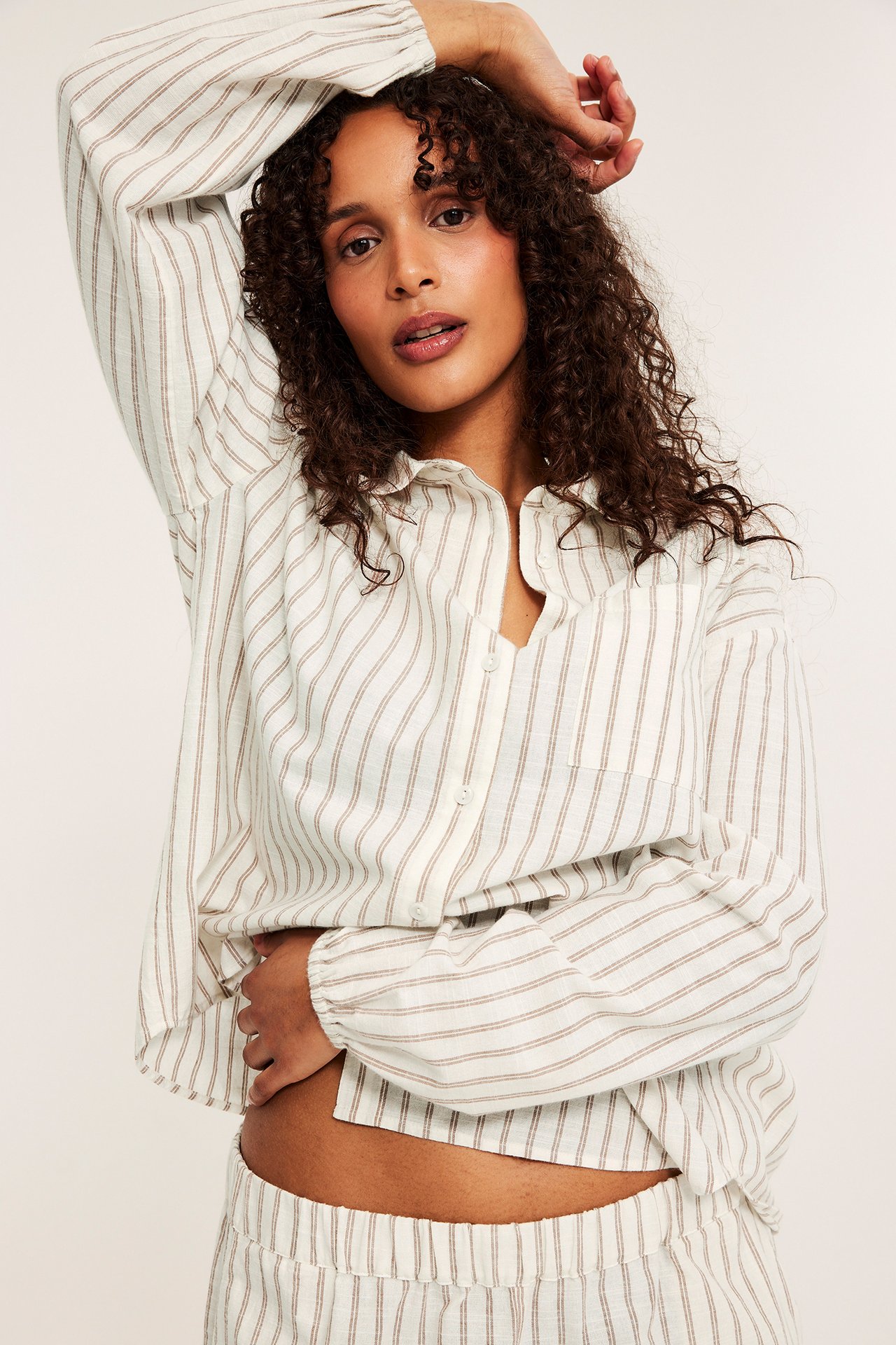 Etunäkymä: Nainen yllään beige raidallinen puuvillapyjamasetti: kauluspaita ja shortsit.