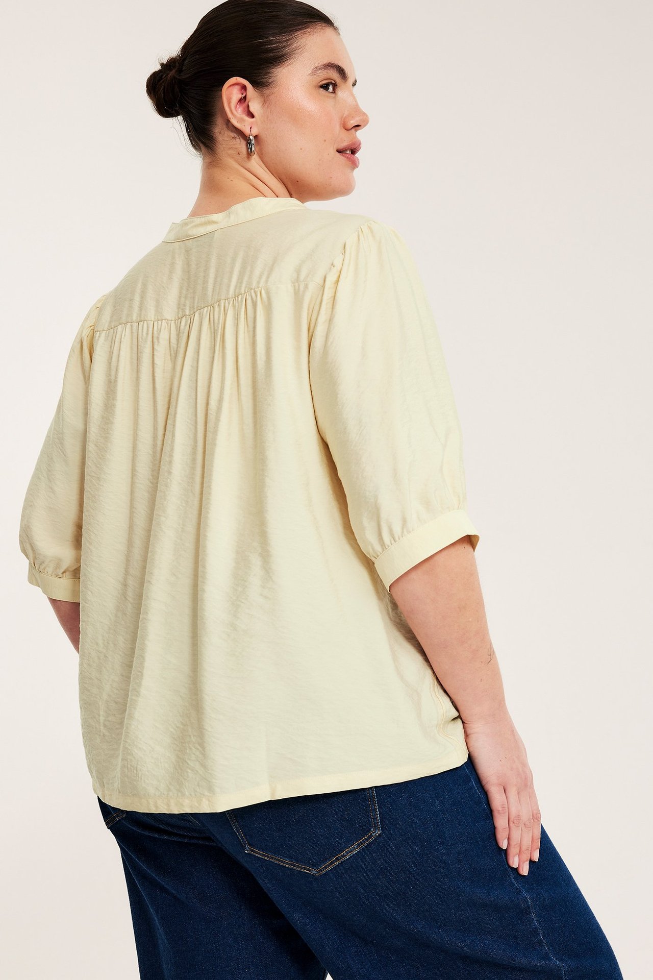 Widok z tyłu kobiety w jasnożółtej bluzce pop-over, z marszczonym tyłem, rękawami 3/4 i ciemnoniebieskich dżinsach.