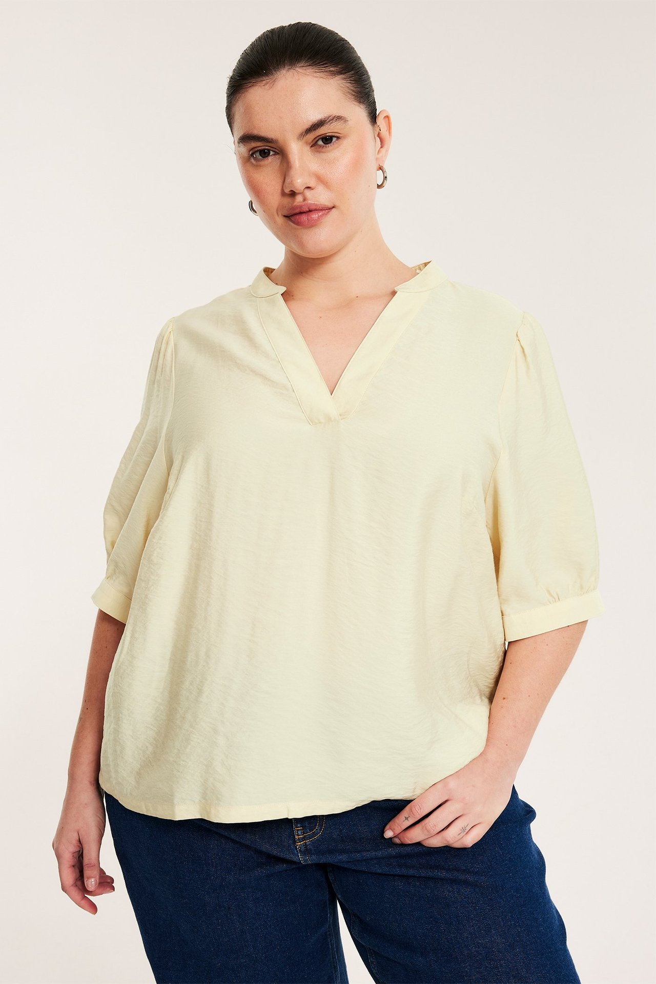 Framifrån: Kvinna i ljusgul pop-over blus med v-ringning, 3/4 puffärm och mörkblå jeans.
