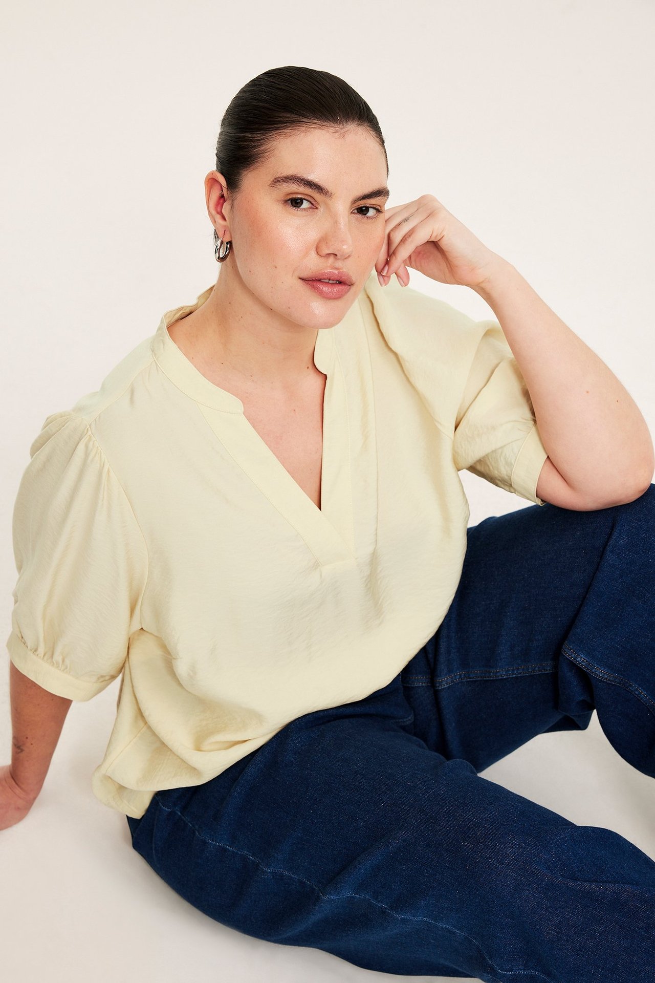 Framfra, kvinne i en lys gul V-hals pop-over-bluse med puffermer og mørkeblå jeans.