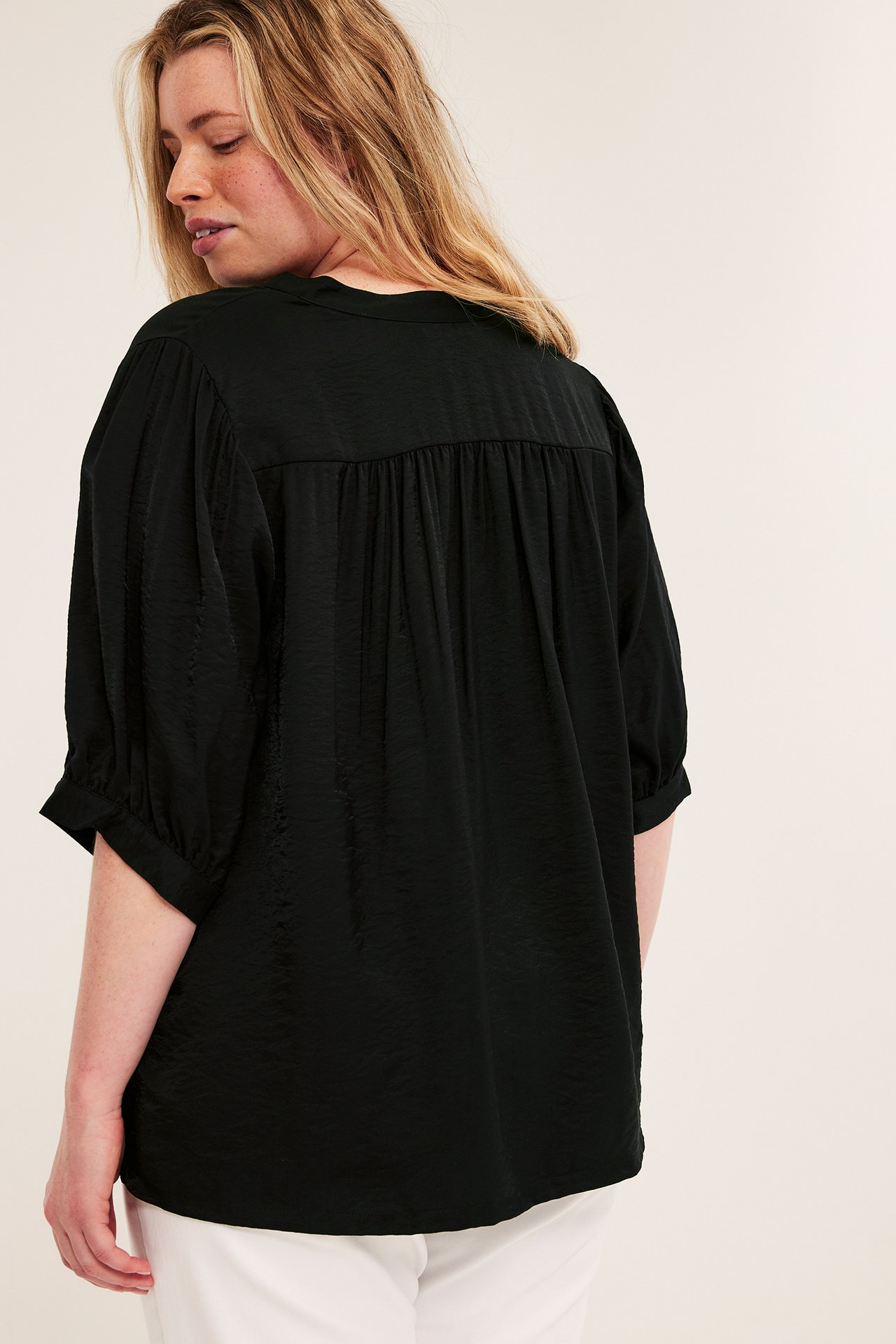 Bakre-sidovy: Kvinna i svart blus med puffärmar, rynkat ok bak och vita byxor.