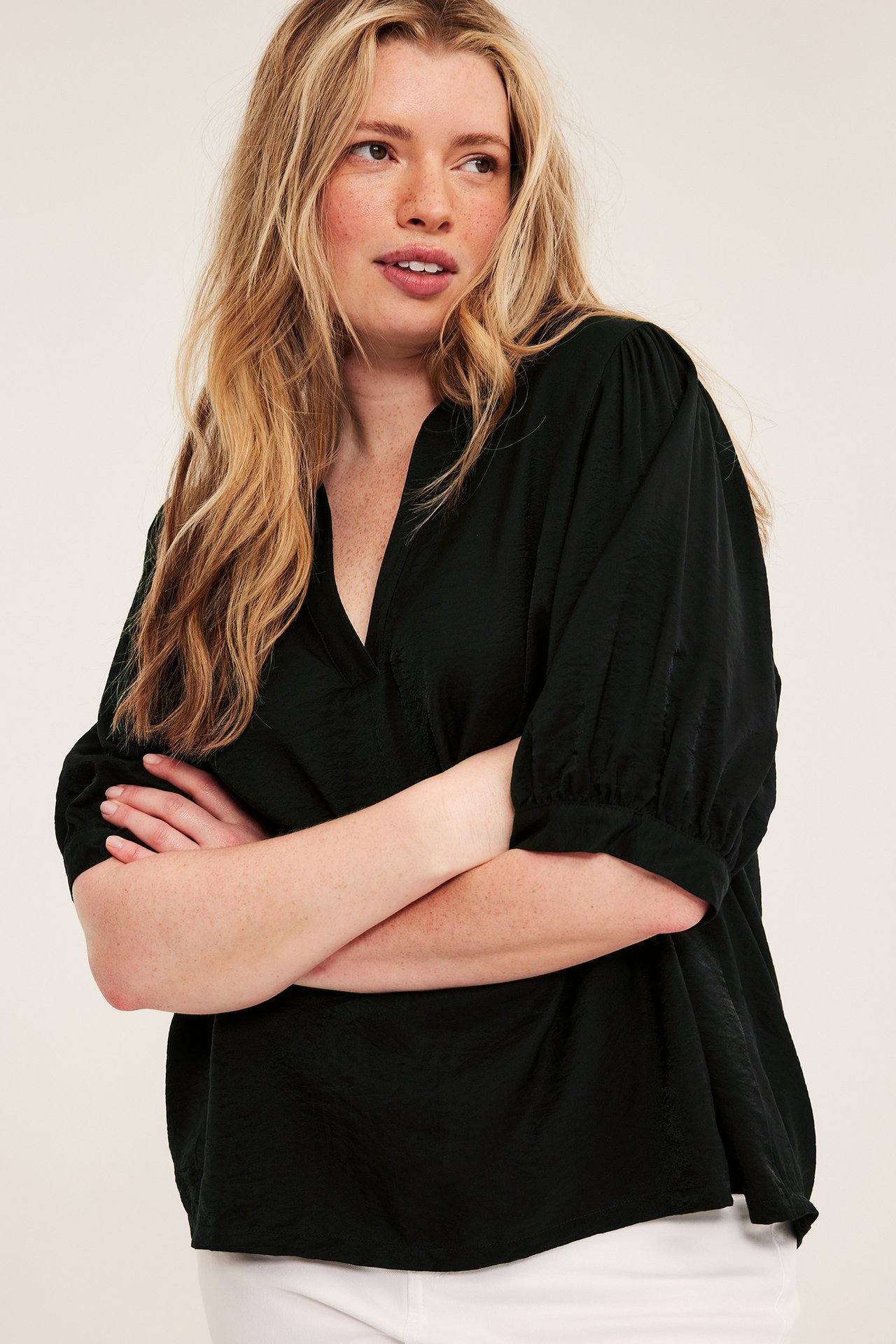 Från sidan: Kvinna i svart V-ringad pop-over-blus med 3/4 puffärmar, över vita byxor.