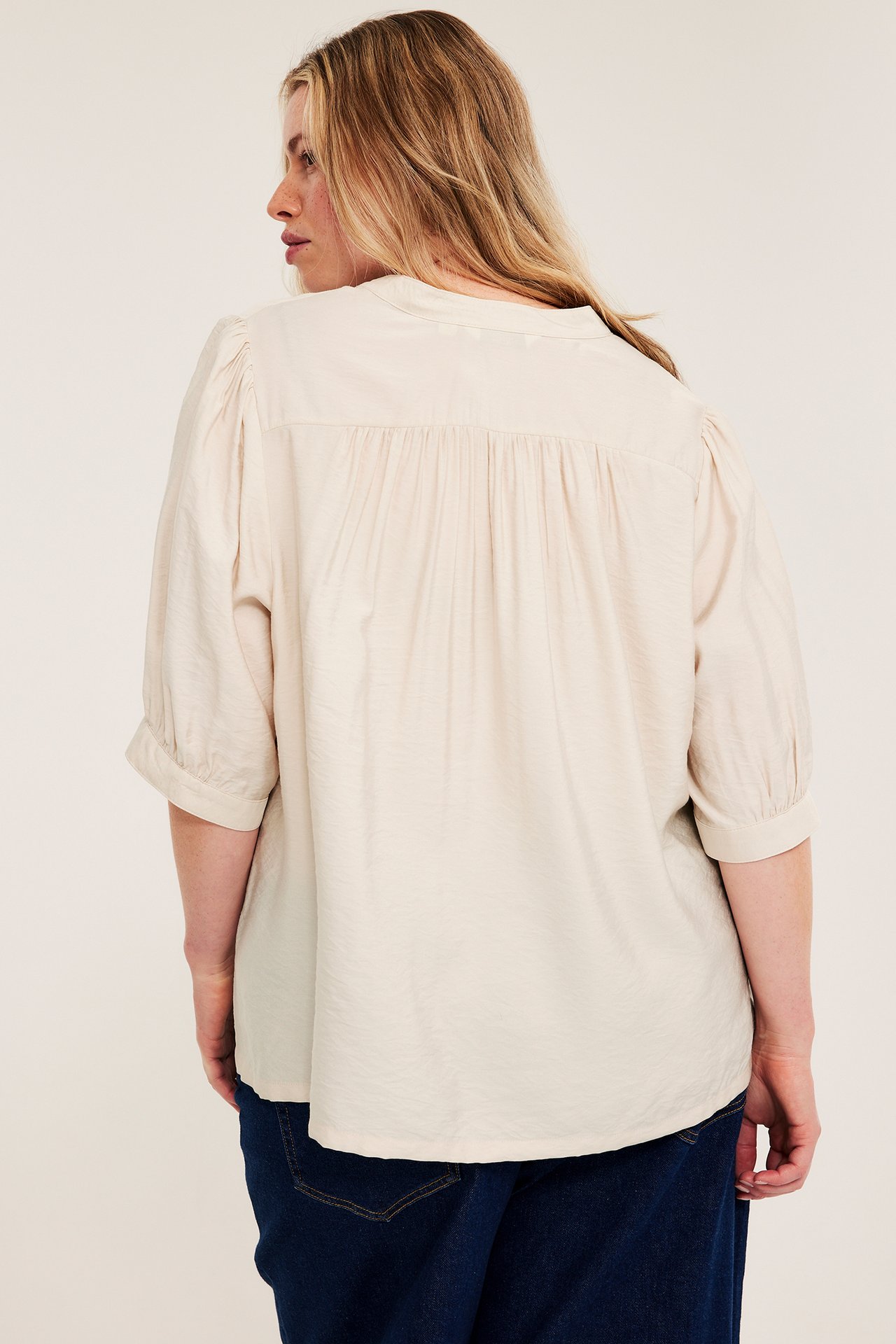 Ryggvy av kvinna i ljusbeige blus med 3/4-puffärmar, veckat ok bak, över mörkblå jeans.