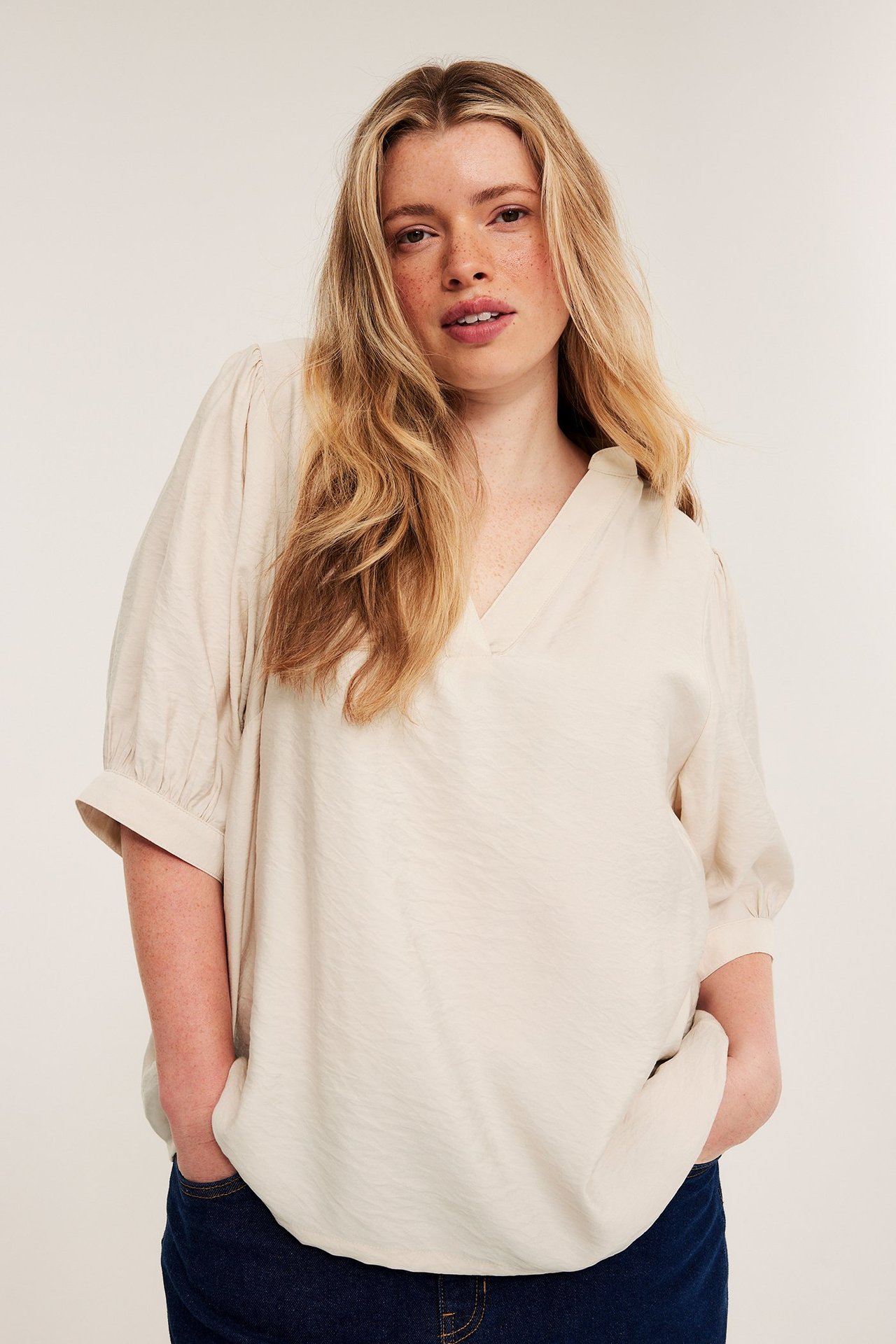 Forfra, kvinne i lys beige V-hals pop-over bluse med 3/4 puffermer og mørkeblå jeans.