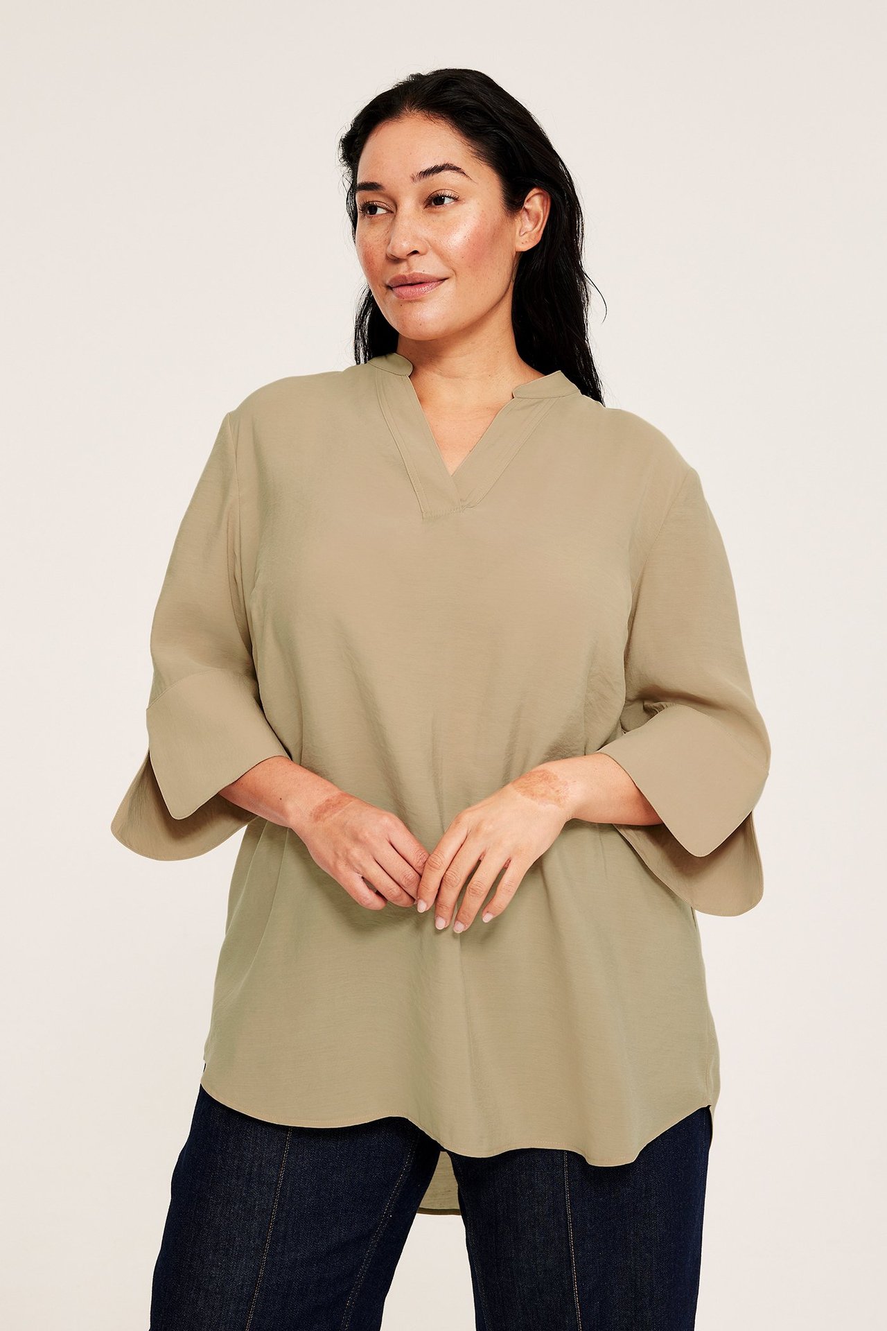 Widok z przodu: kobieta w luźnej beżowej bluzce z dekoltem w serek, rozszerzanymi rękawami 3/4 i ciemnymi dżinsami.