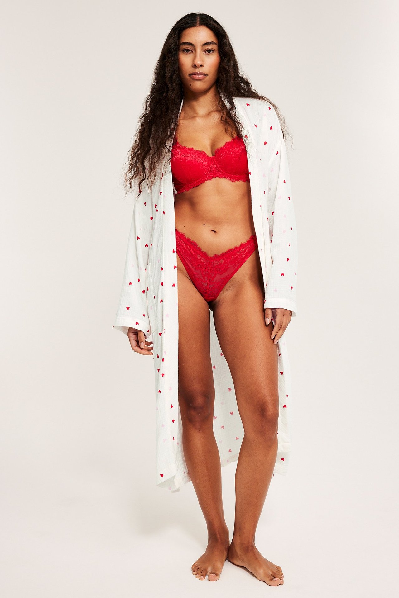 Forfra: kvinne i åpen offwhite bomullsmorgenkåpe med hjerter, over rød blonde-BH og truse.