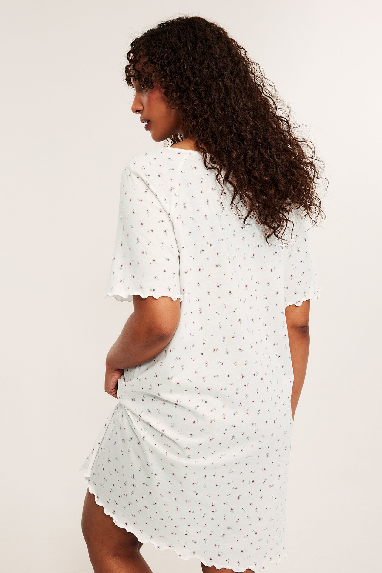 Bakfra, dame i lysmønstret nattkjole i bomullsjersey med rosa blomstermønster og bølgede kanter.