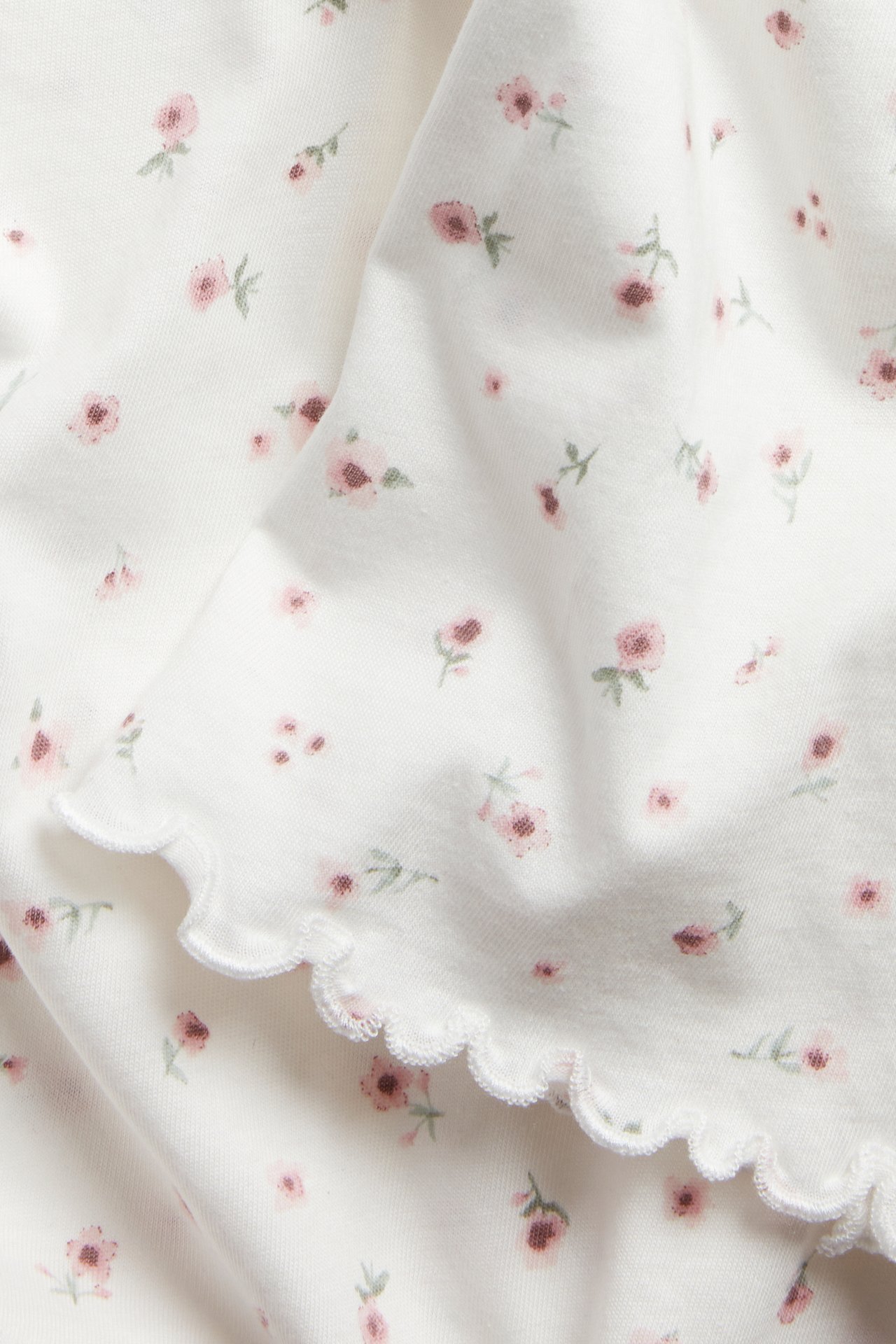 Lähikuva valkoisesta puuvillaisesta yöpaidan kankaasta, jossa vaaleanpunainen kukkakuvio ja aaltoileva reuna.
