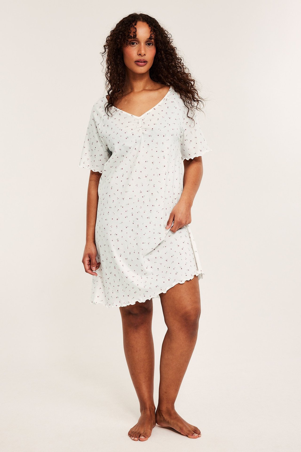 Frontvy av en kvinna i ett vitt bomullsjersey nattlinne med rosa blommigt mönster och vågiga kanter.