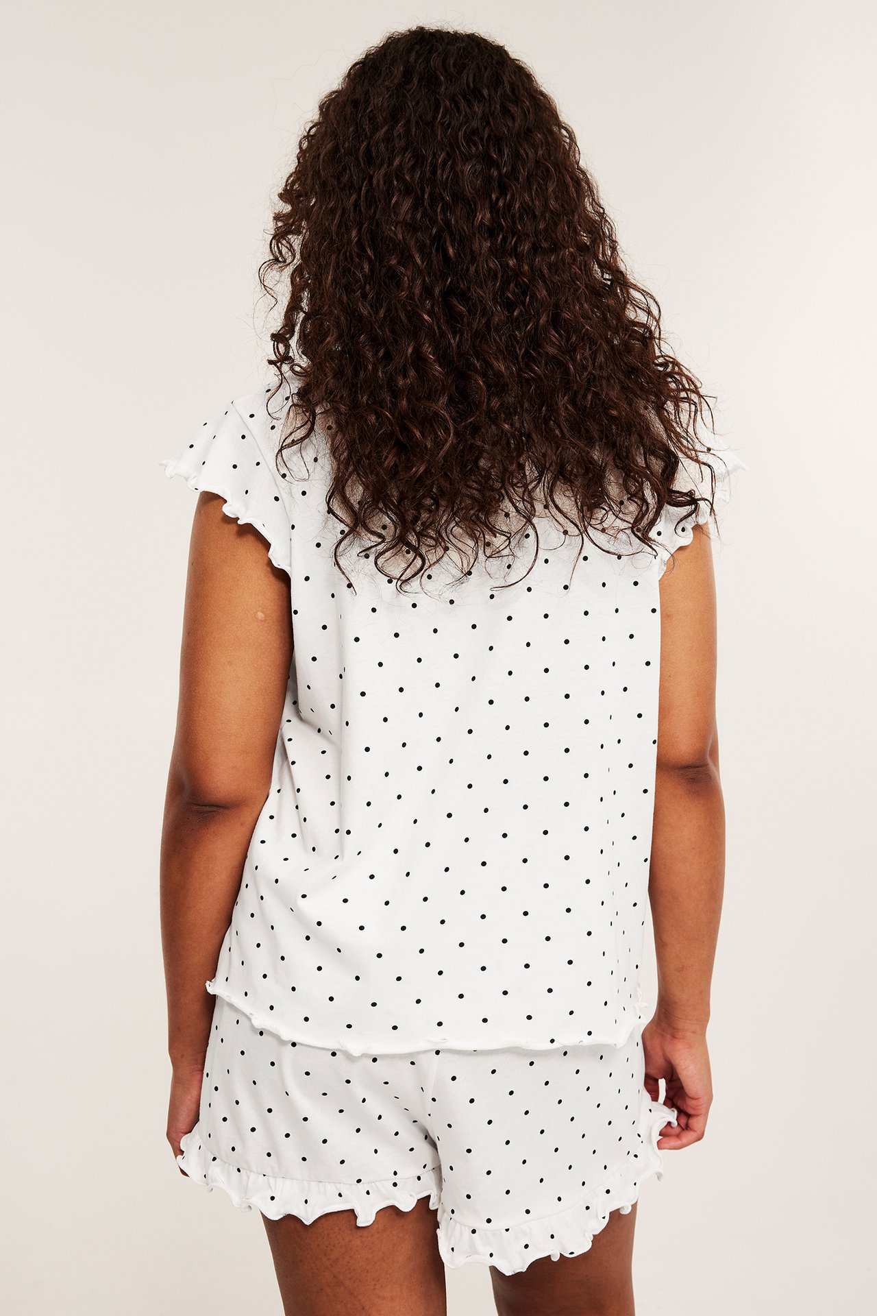 Naisen selkäpuoli: luonnonvalkoinen pilkullinen puuvillapyjama röyhelötopilla ja röyhelöshortseilla.