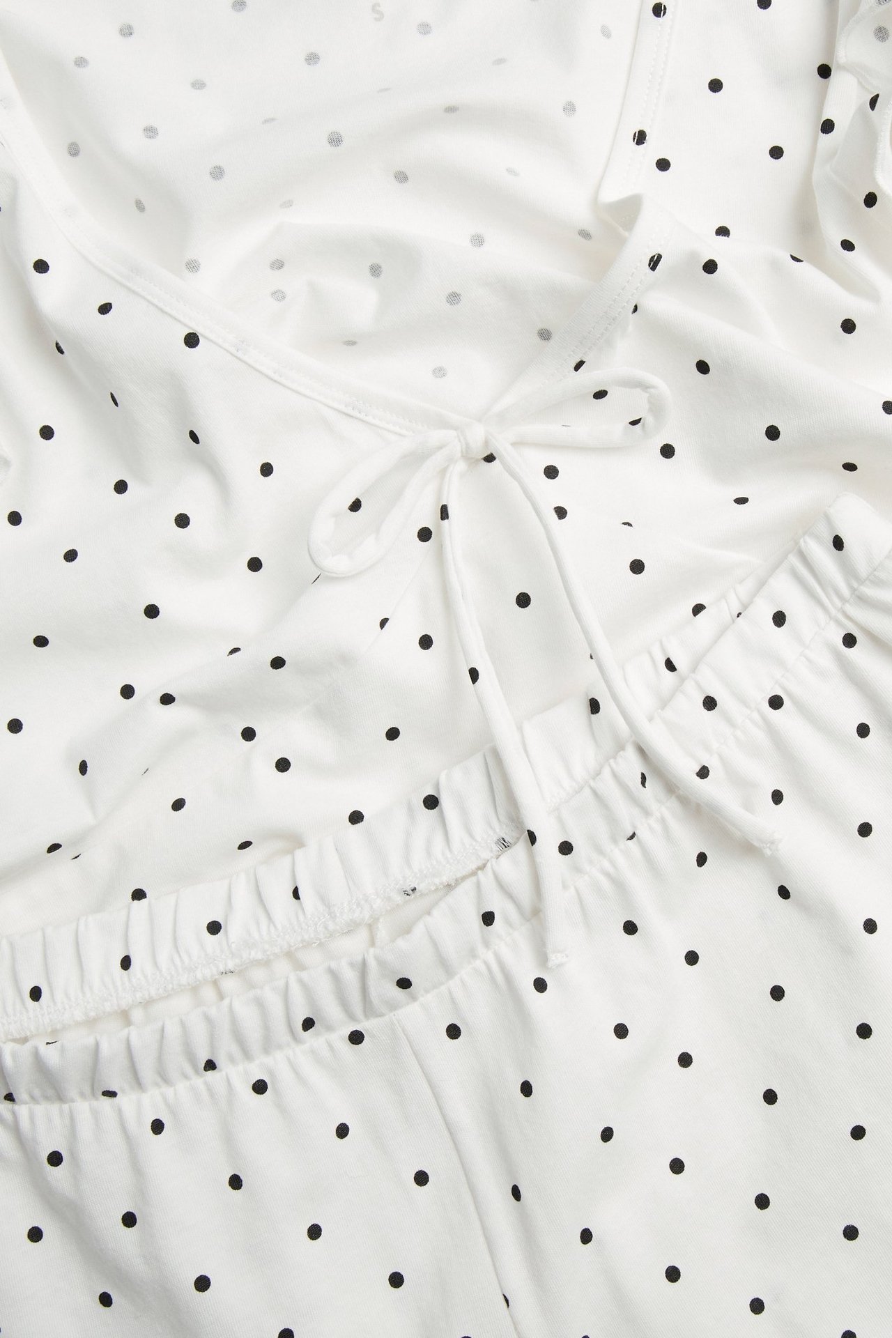 Lähikuva luonnonvalkoisesta pilkullisesta trikoopyjamasta: V-aukkoinen toppi rusetilla, shortsit joustavalla vyötäröllä.