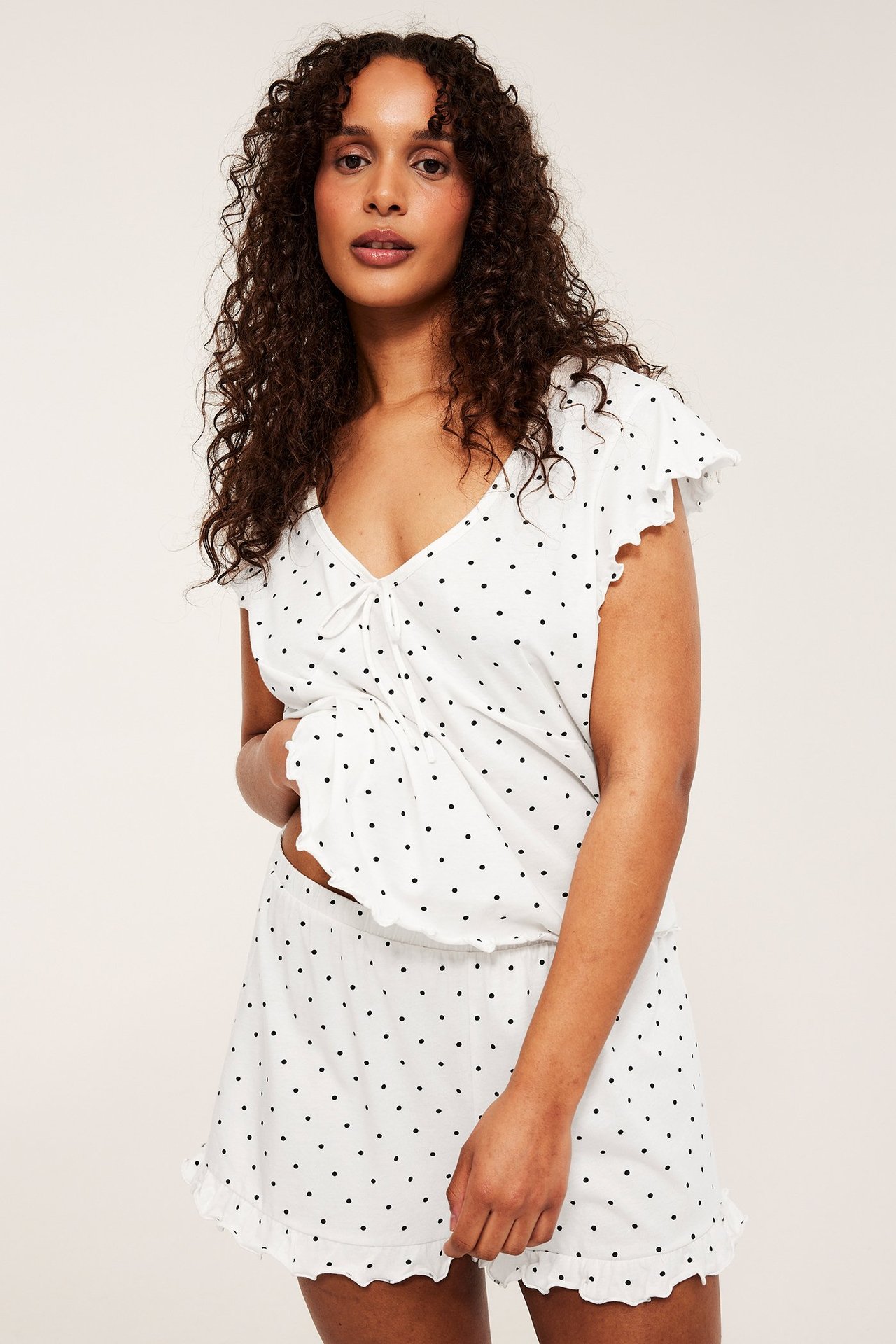 Widok z przodu: kobieta w kremowej piżamie w kropki z bawełny, top z dekoltem V i szorty z falbanką.