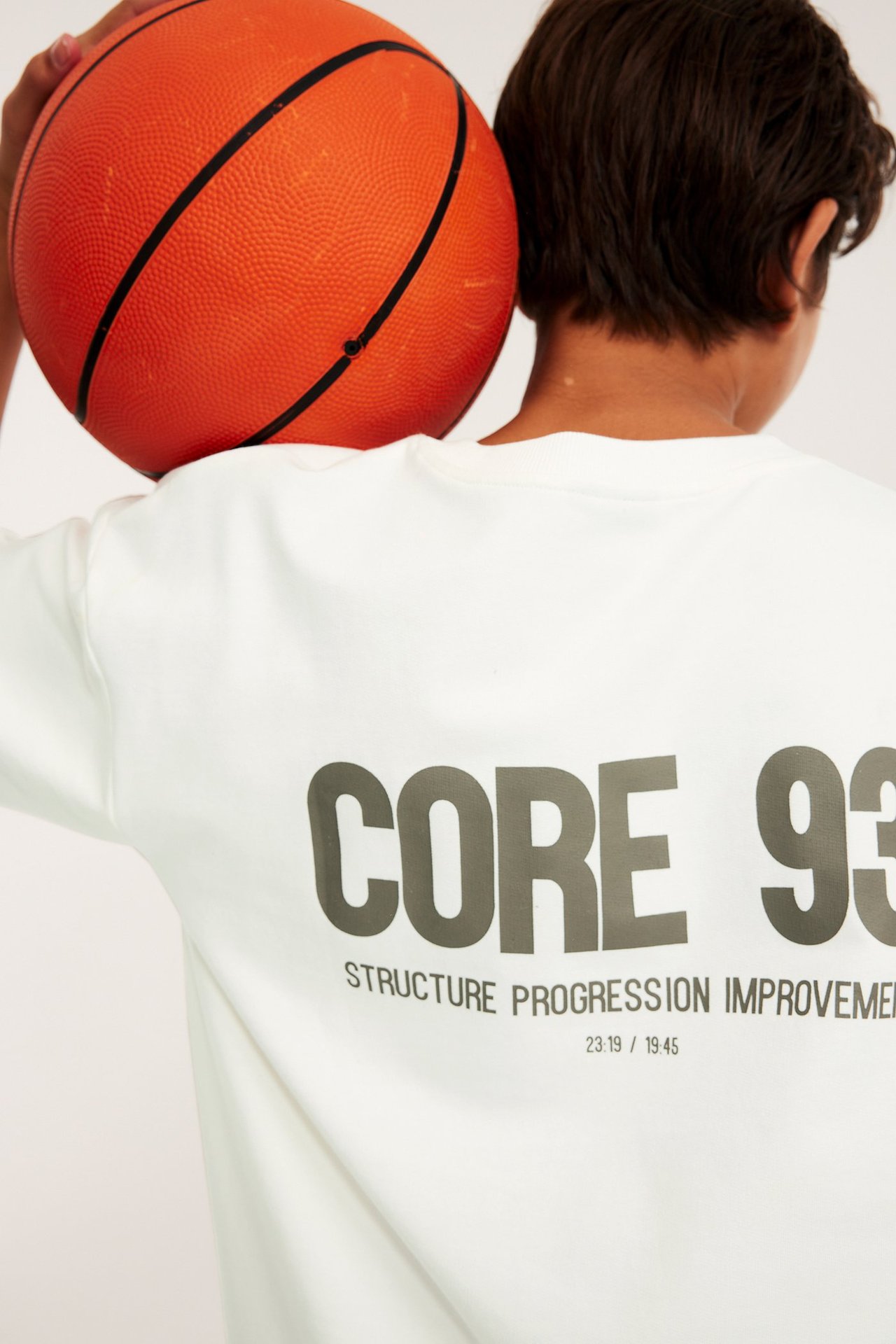 Bakfra: barn i en hvit oversized bomulls-t-skjorte med grått ryggtrykk, holder en oransje basketball.