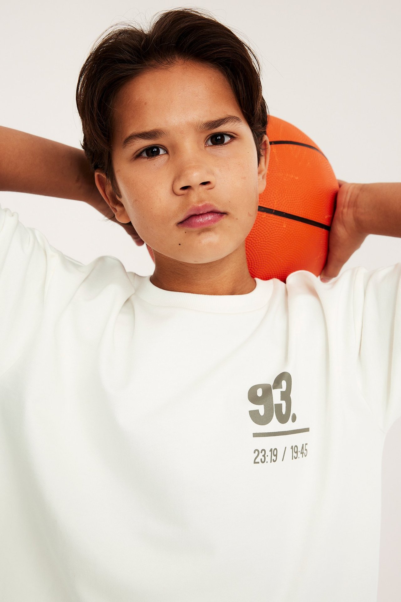Nærbilde forfra: barn bærer hvit oversized bomulls-t-skjorte med fronttrykk, holder en oransje basketball.