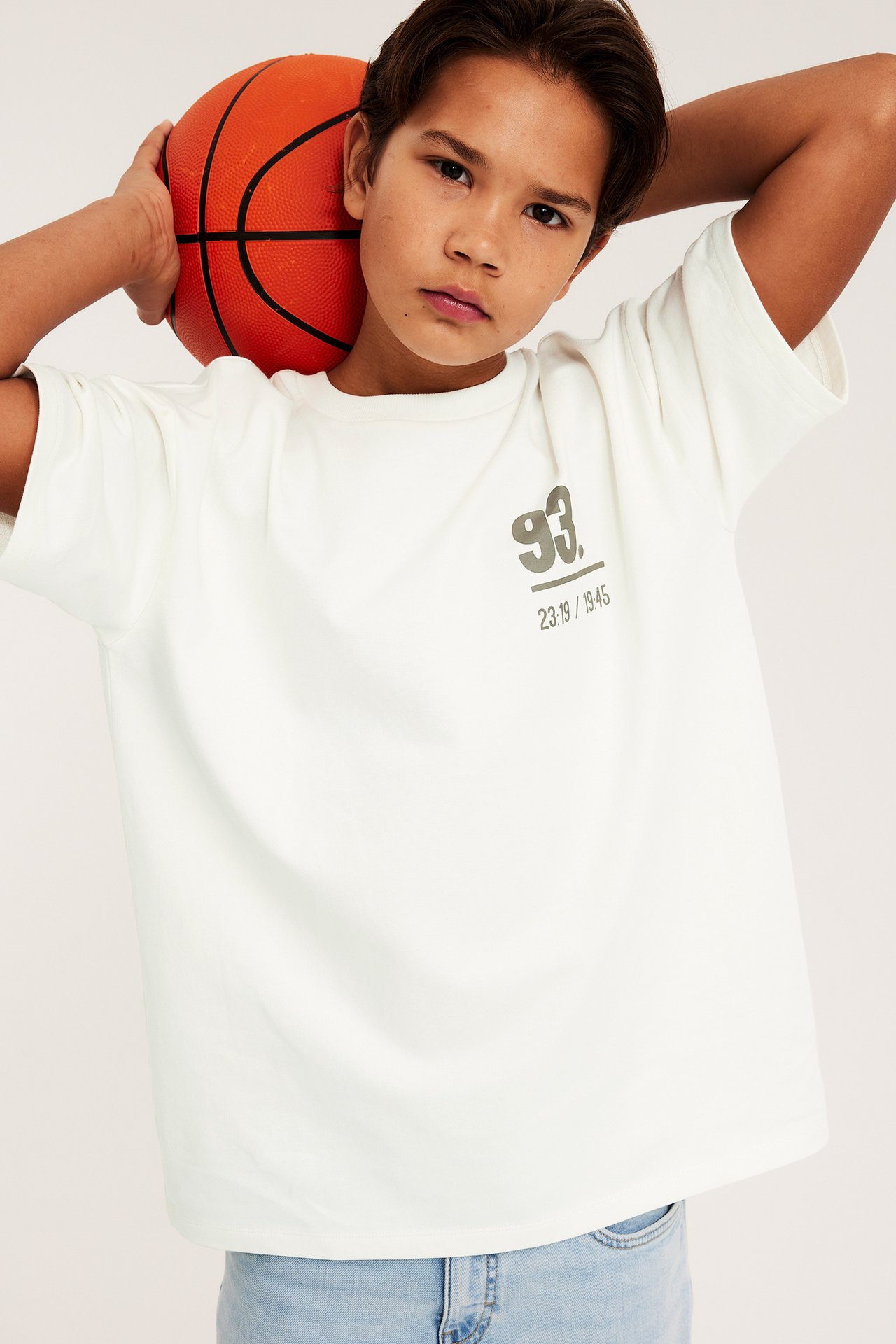 Widok z przodu: Dziecko w białym obszernym t-shircie z szarym nadrukiem, jasnoniebieskich dżinsach, z pomarańczową piłką.