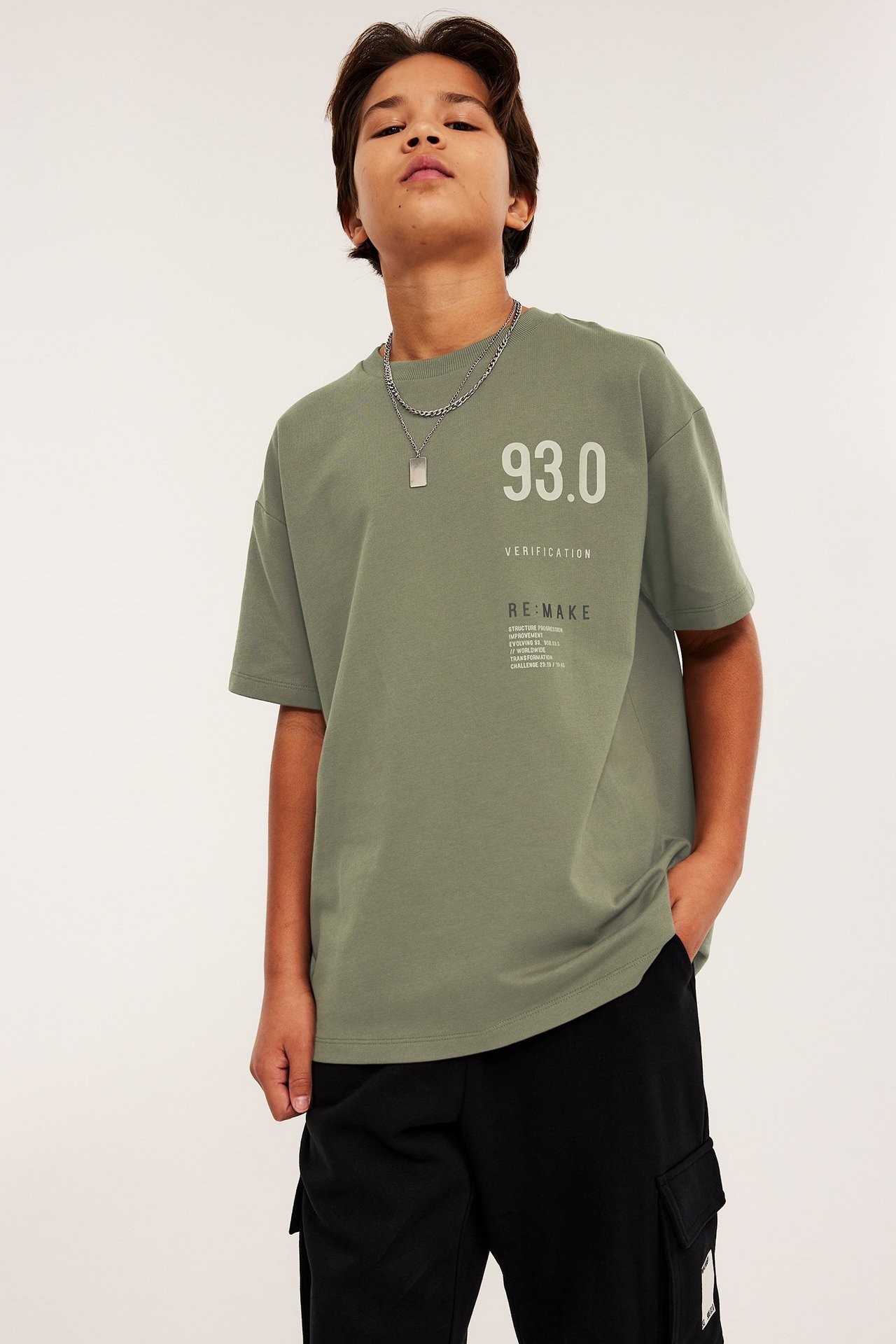 Edestä poika yllään oversized vihreä puuvillainen t-paita painatuksella, mustat housut ja hopeakaulakoru.
