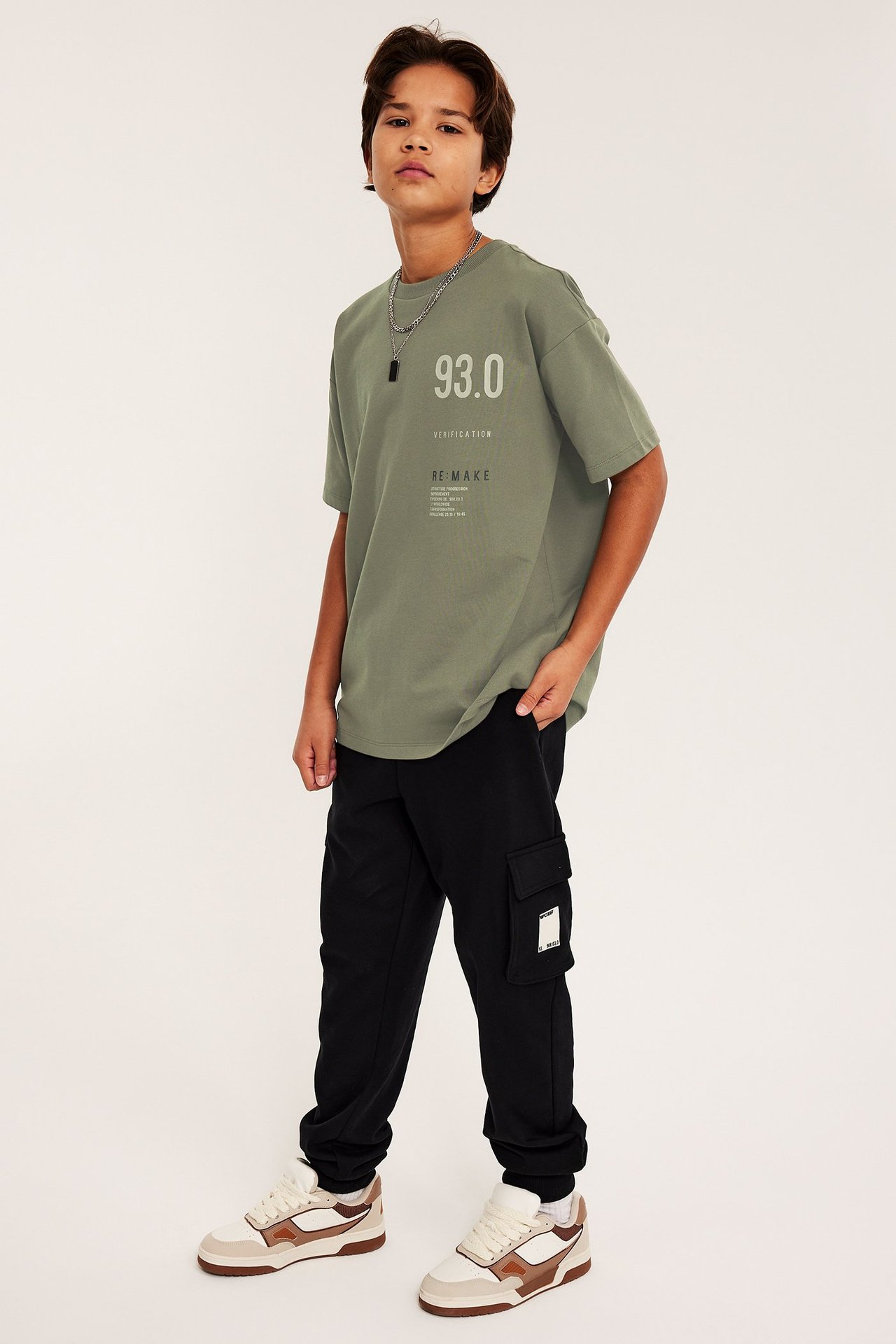 Edestä: Lapsi vihreässä painetussa oversized T-paidassa, mustissa cargo-housuissa ja kaksisävyisissä tennareissa.