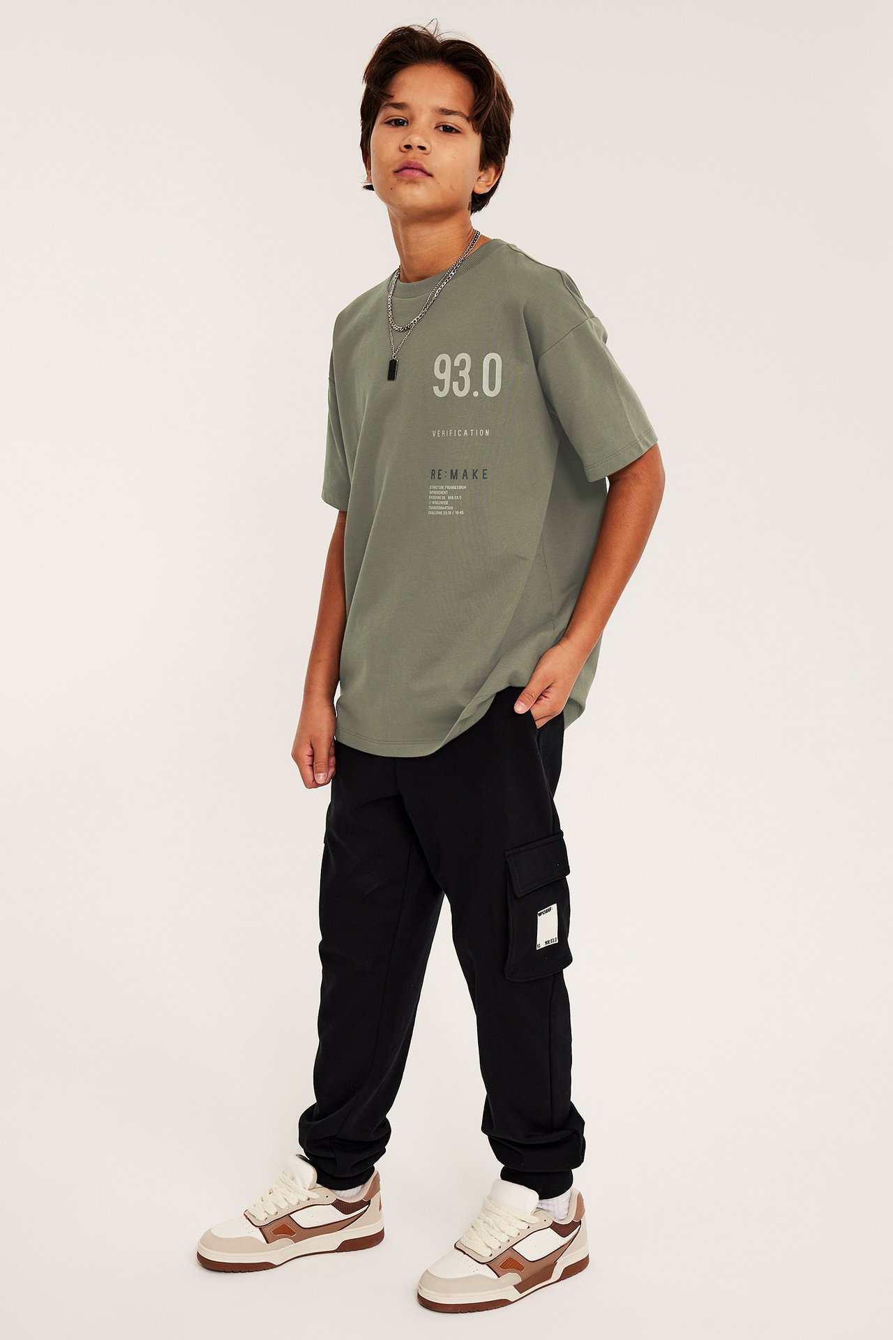 Widok z przodu dziecka w zielonej, obszernej bawełnianej koszulce z nadrukiem na piersi, czarnych spodniach cargo i sneakersach.