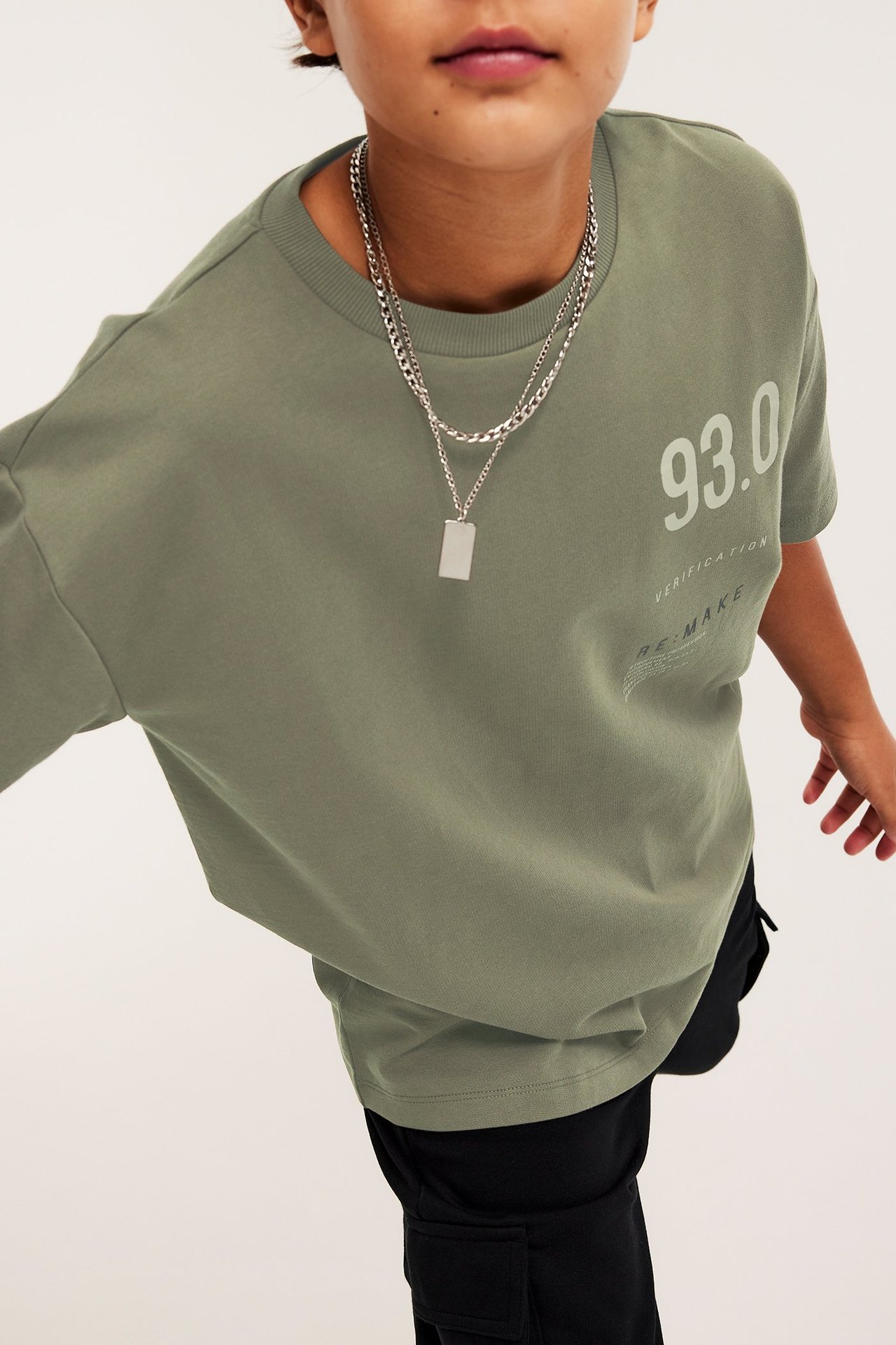 Etunäkymä: Lapsi yllään vihreä oversized puuvilla-T-paita painatuksella, mustat cargohousut ja kerrostetut hopeakaulakorut.