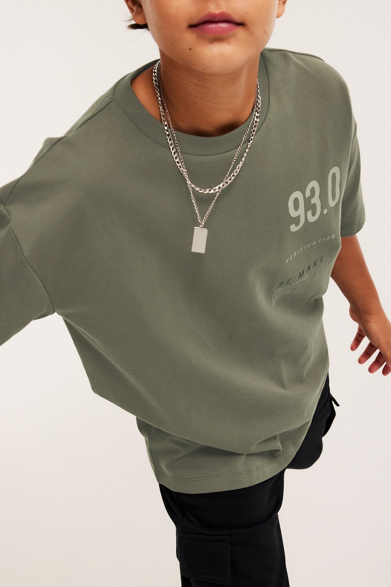 Dziecko nosi zielony bawełniany t-shirt oversize z nadrukiem, czarne spodnie, srebrne naszyjniki.