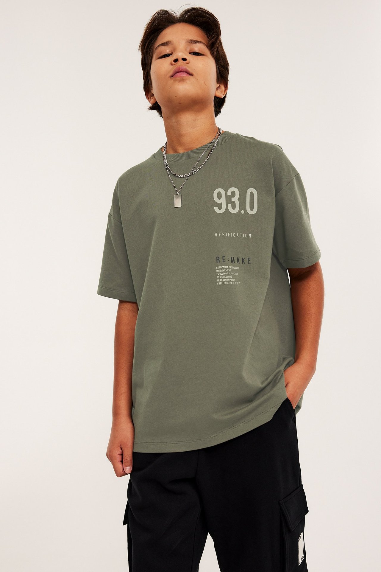 Edestäpäin, lapsi yllään vihreä oversized puuvilla t-paita rintapainatuksella ja mustat cargohousut.