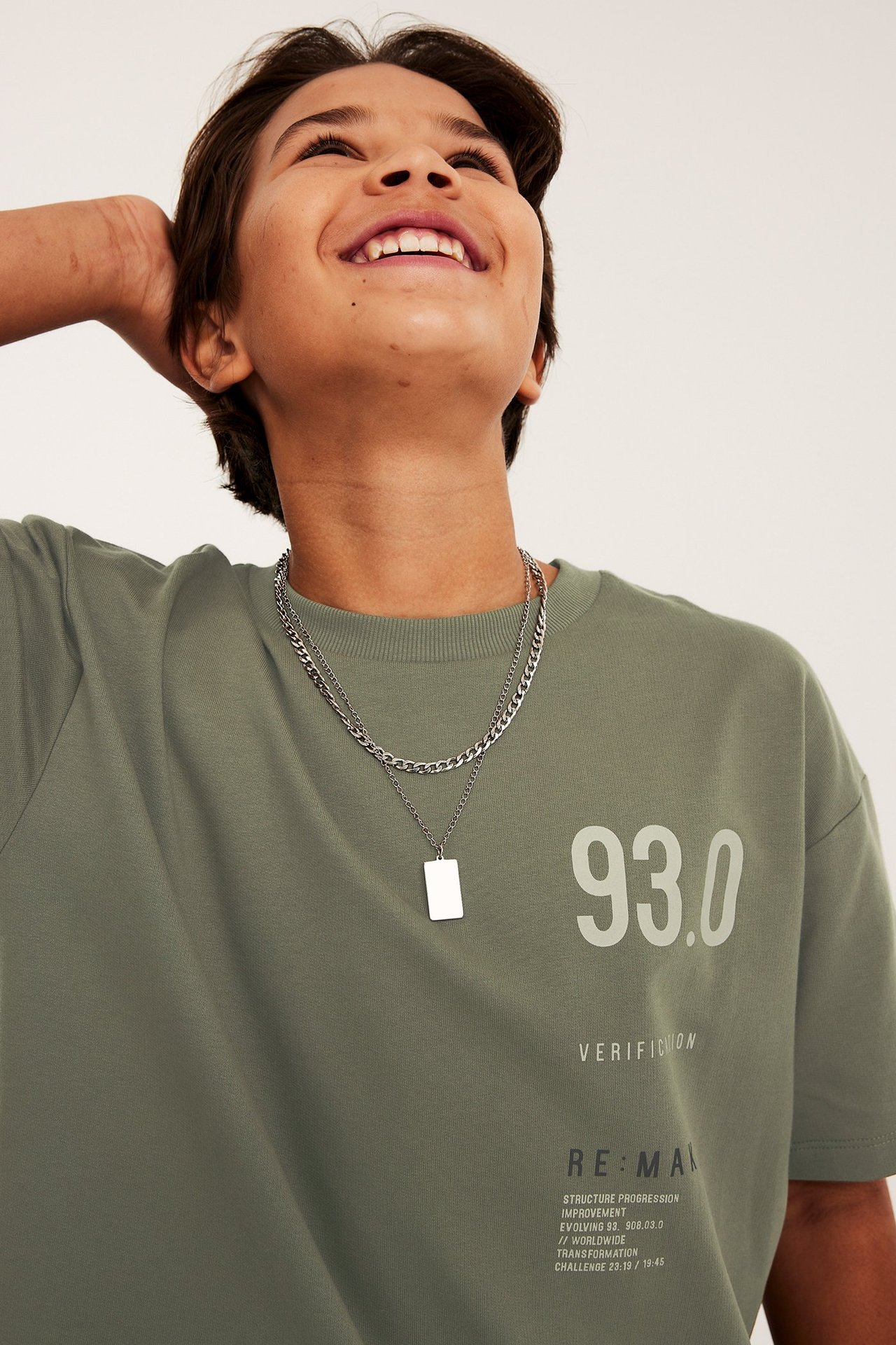 Lähikuva edestä: lapsi yllään ylisuuri vihreä puuvilla-T-paita rintapainatuksella ja hopeinen kerroskaulakoru.