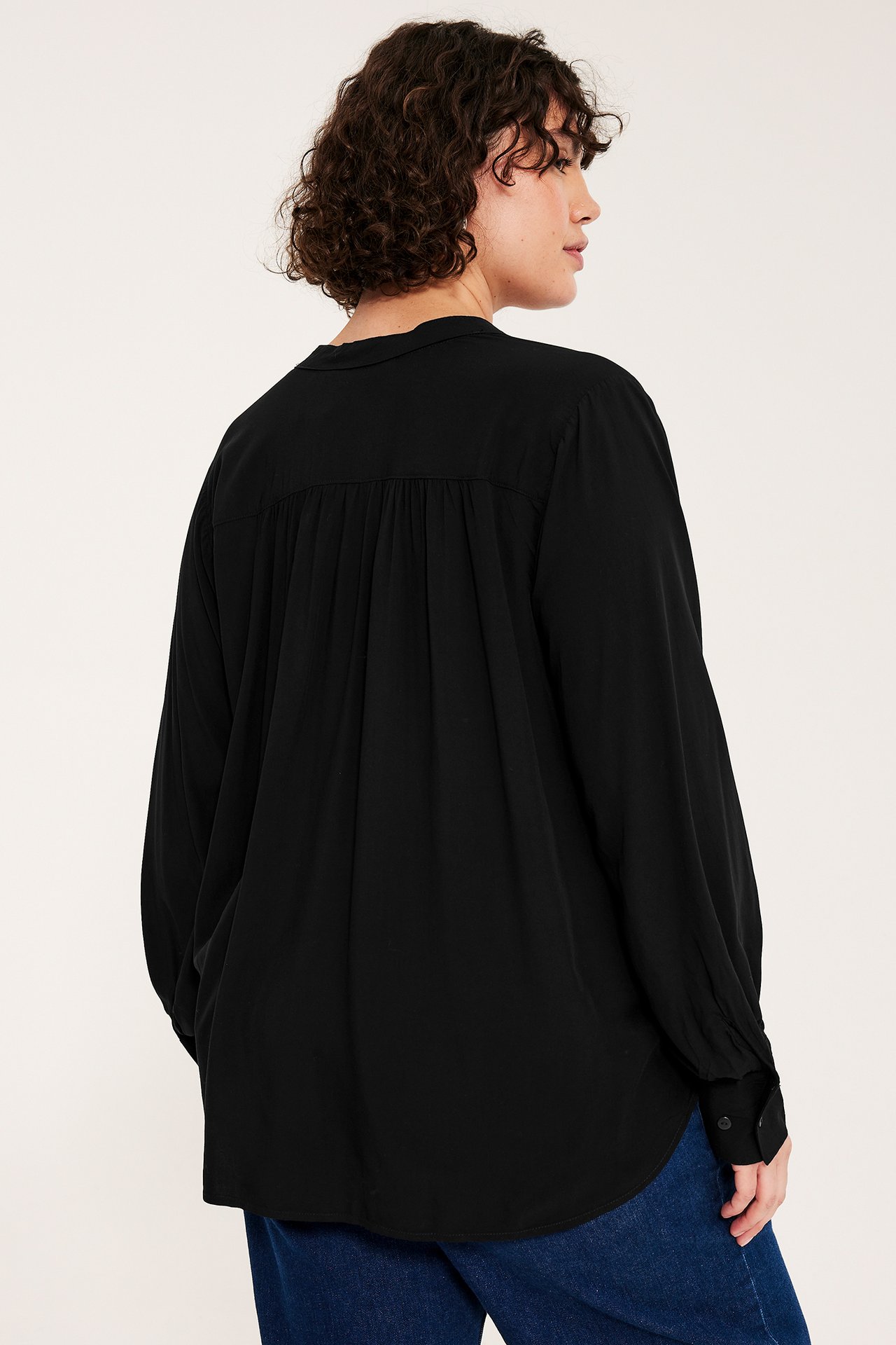 Widok z tyłu kobiety w czarnej bluzce wiskozowej z dekoltem V, bufiastymi rękawami i niebieskich dżinsach.