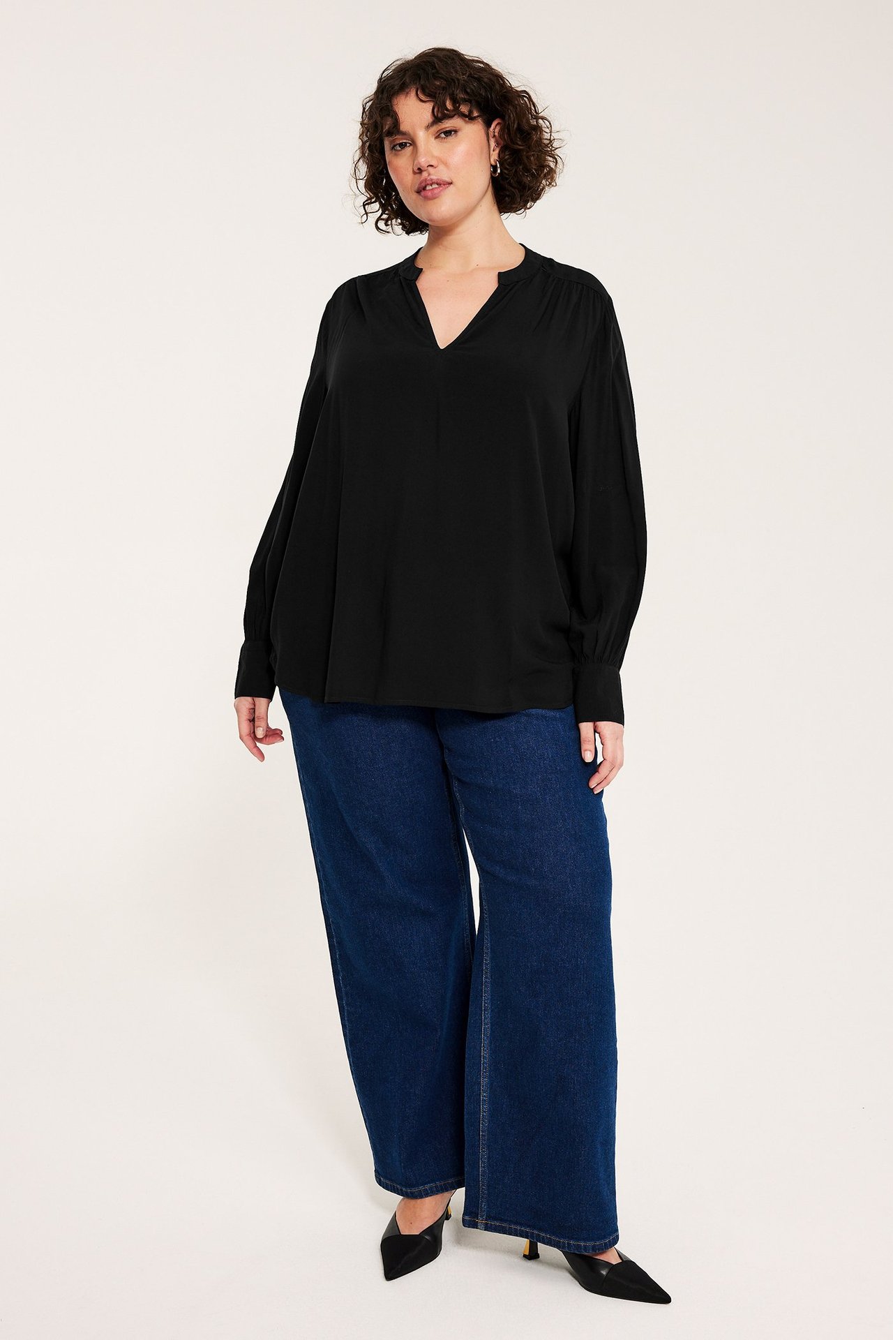 Widok z przodu: Kobieta w czarnej bluzce wiskozowej V-neck, bufiastymi rękawami, ciemnoniebieskich dżinsach i czarnych szpilkach.