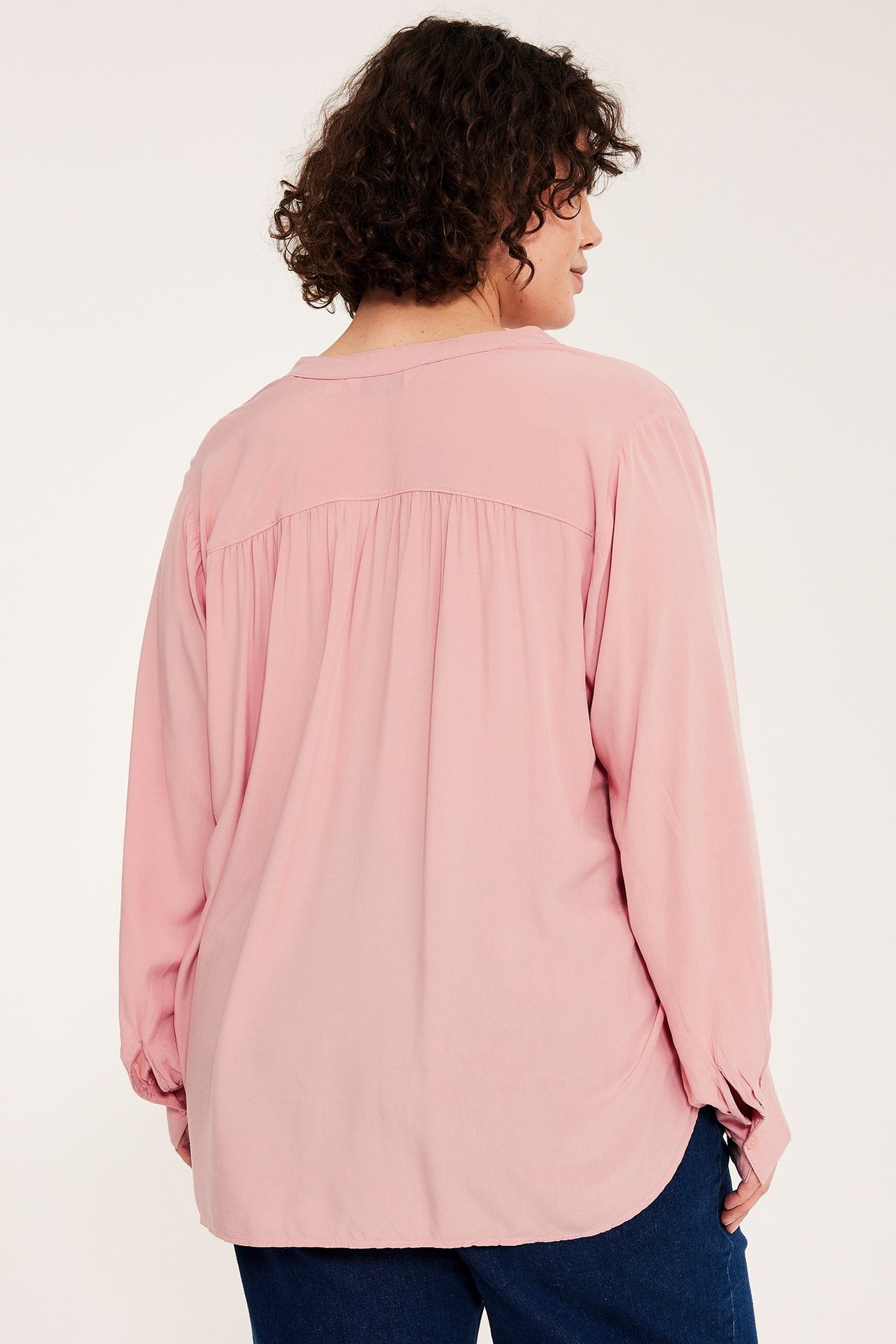 Nainen takaapäin, yllään väljä vaaleanpunainen viskoosipusero, takana poimutettu kaarroke ja tummansiniset housut.