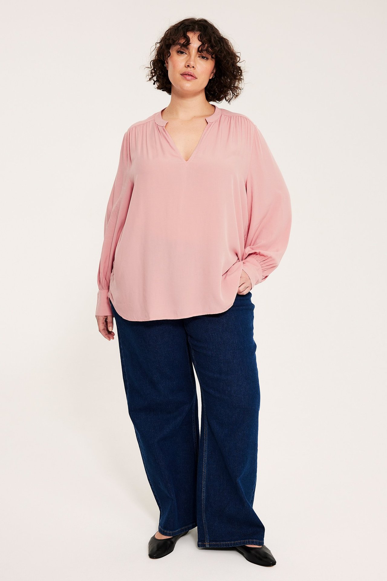 Etunäkymä: Nainen pinkissä V-pääntie viskoosipuserossa, pussihioilla, tummansinisissä farkuissa ja mustissa kengissä.