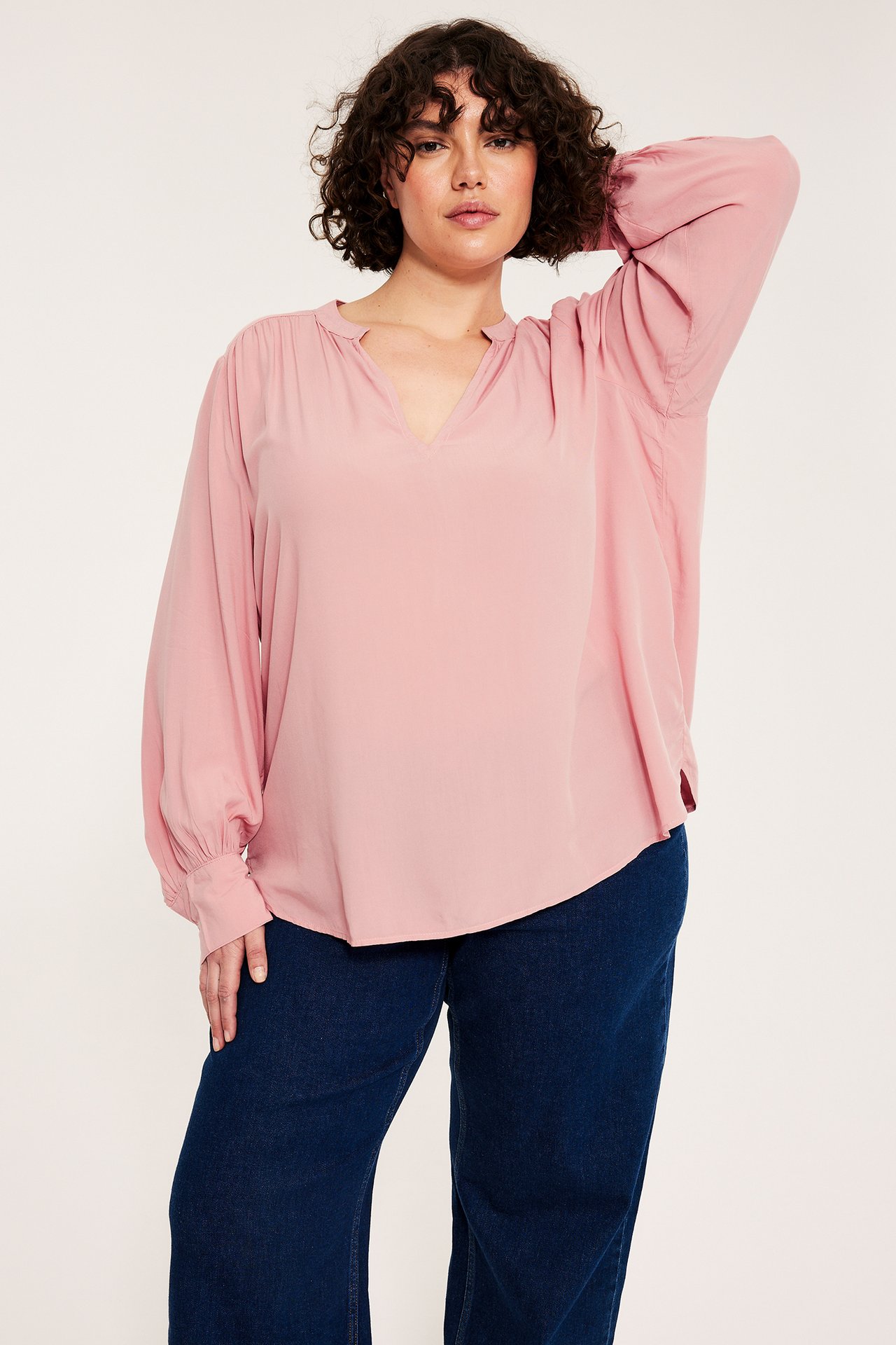 Naisen etunäkymä, yllään vaaleanpunainen V-kaula-aukollinen viskoosipusero pussihioilla ja tummansiniset housut.